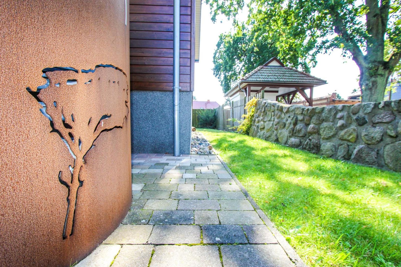 Pflasterweg neben Hauswand mit Metallbaum. Grasfläche und Steinmauer im Hintergrund.