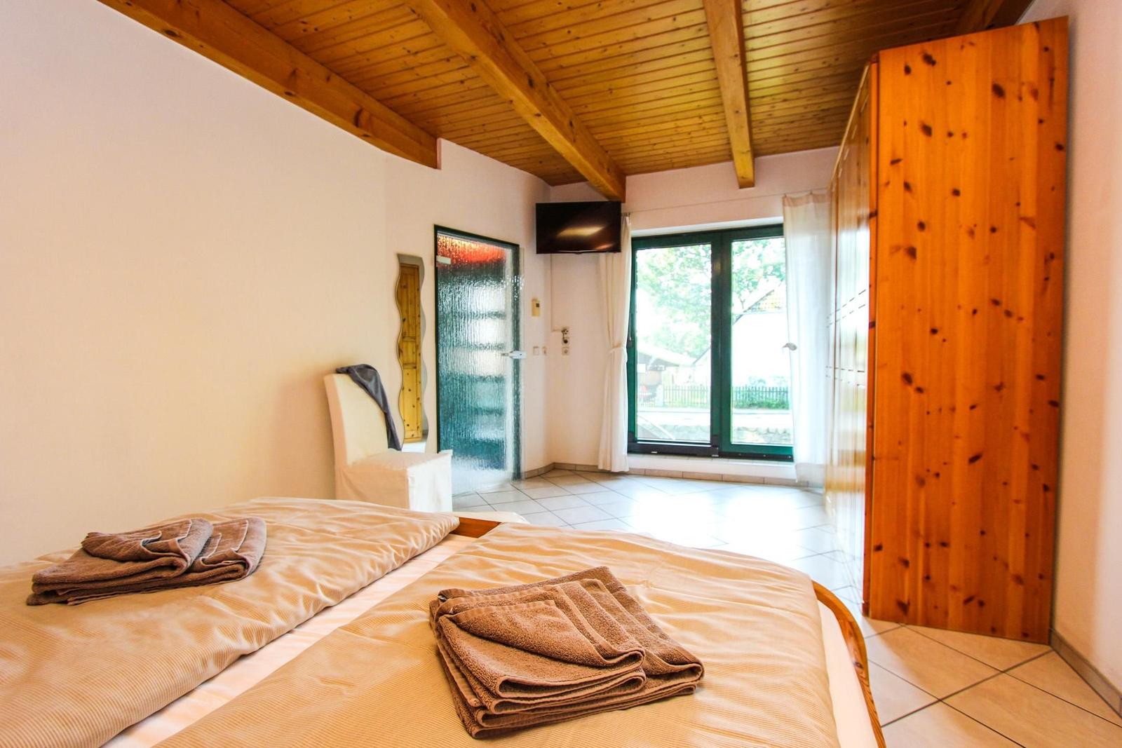 Schlafzimmer mit Bett, Holzbalkendecke und Fenster. Holzwand und TV sichtbar.