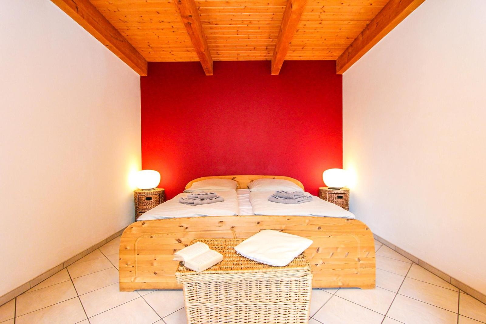 Schlafzimmer mit Holzbett, roter Wand und Holzdecke. Beleuchtung und Körbe sind vorhanden.