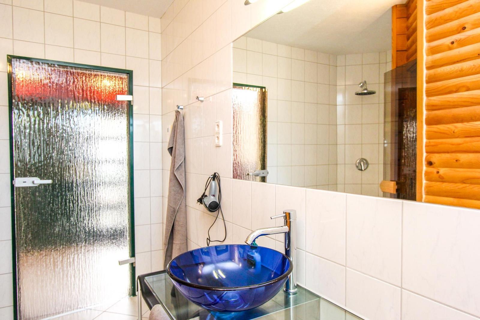 Badezimmer mit blauem Waschbecken, Spiegel und Dusche. Holzwand und gefärbte Tür sichtbar.
