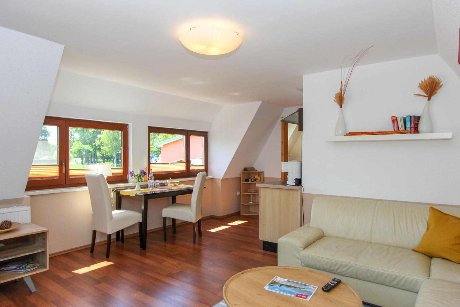 Wohn- und Essbereich mit Sofa, Tisch, Stühlen und Fenstern. Holzboden und weiße Wände.