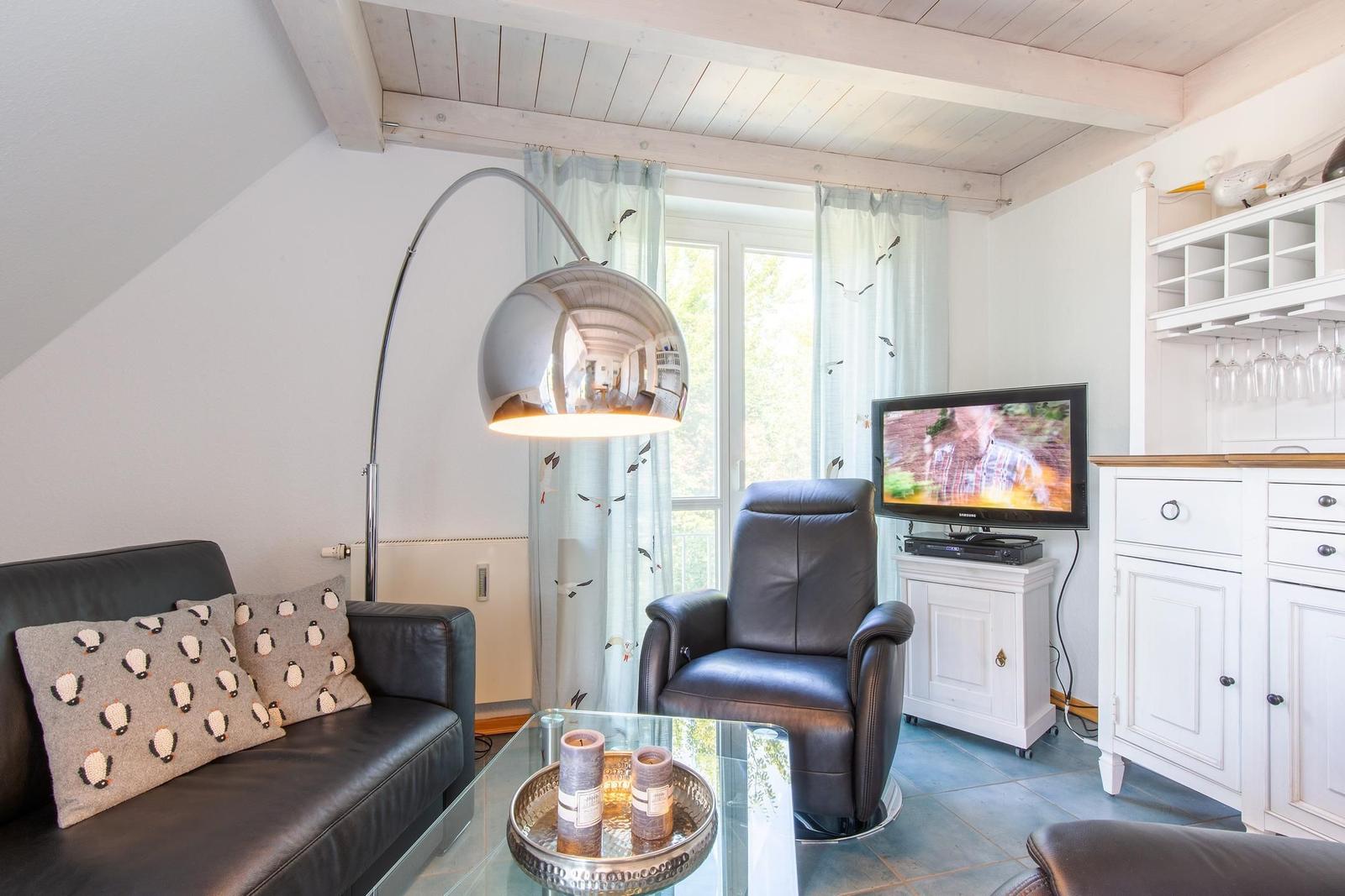 Wohnzimmer mit Sofa, Sessel, TV und Glas-Tisch unter Holzbalkendecke.