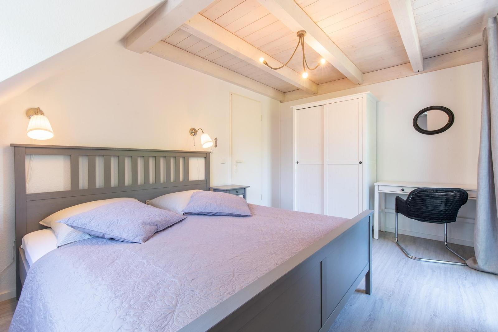 Schlafzimmer mit Bett, Schrank und Schreibtisch unter Holzbalkendecke.