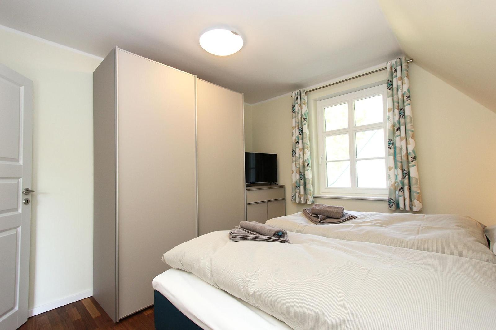 Schlafzimmer mit Doppelbett, Kleiderschrank, TV und Fenster mit Vorhängen.