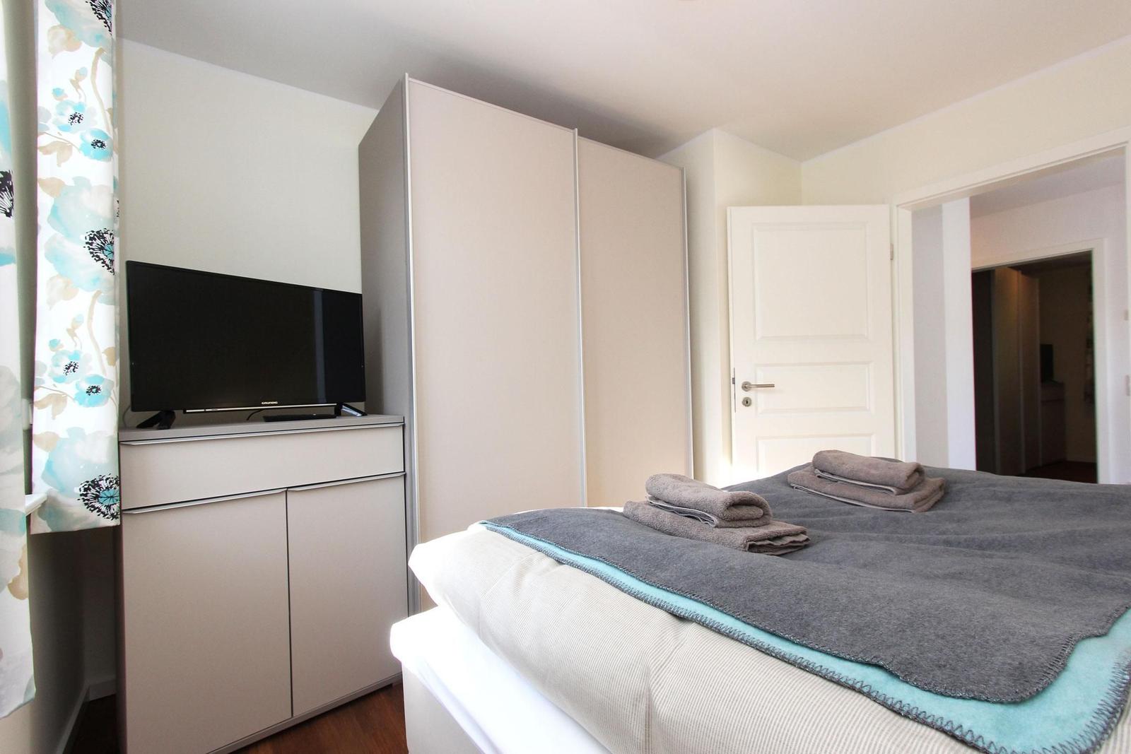 Schlafzimmer mit Bett, Kleiderschrank und Fernseher. Tür führt in angrenzenden Raum.