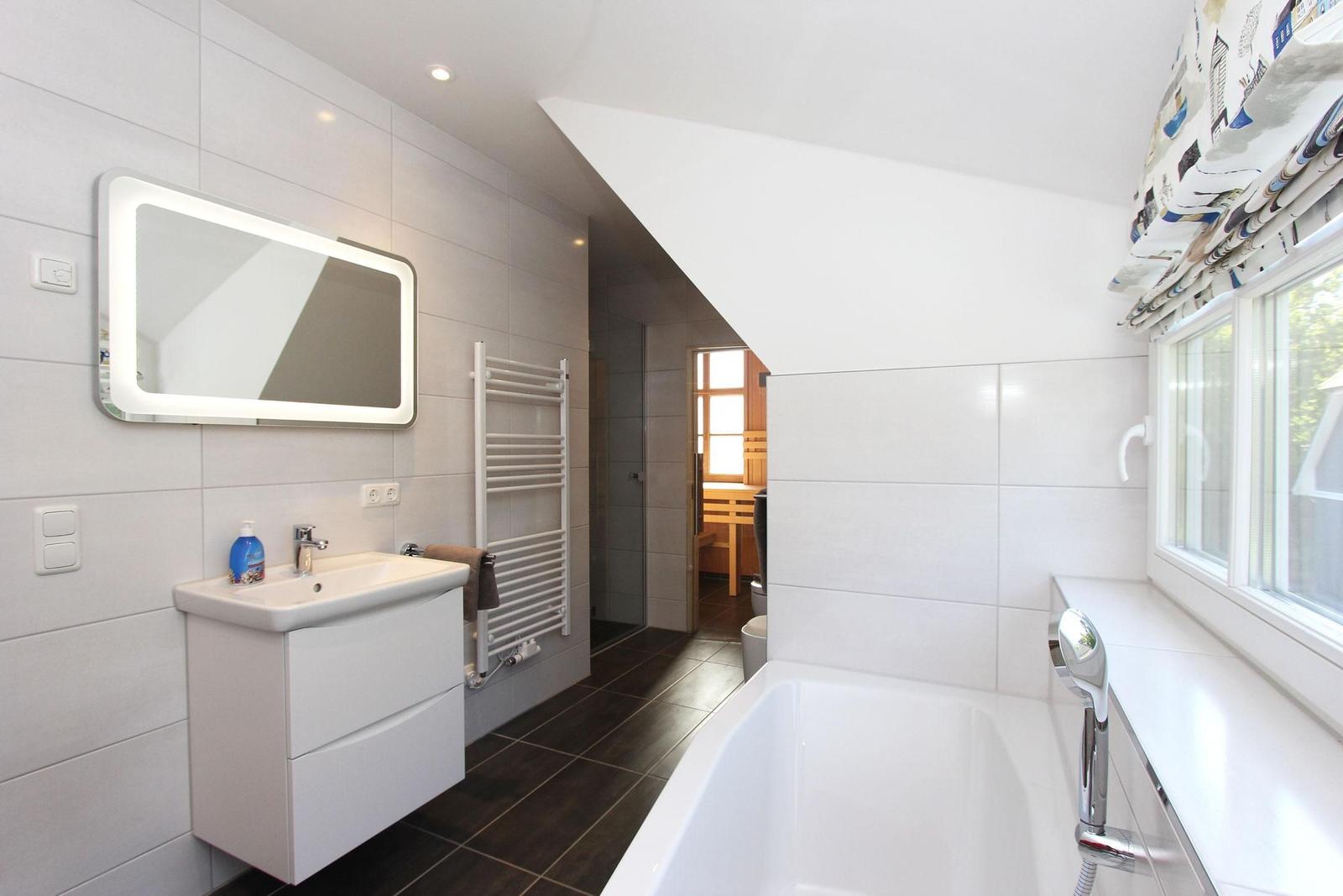 Badezimmer mit Badewanne, Waschbecken und Spiegel. Durchgang zur Sauna sichtbar.