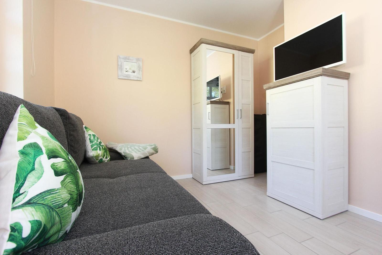 Grauer Sofa mit grün-weißen Kissen, weißer Schrank und Fernseher auf Holzkonsole.