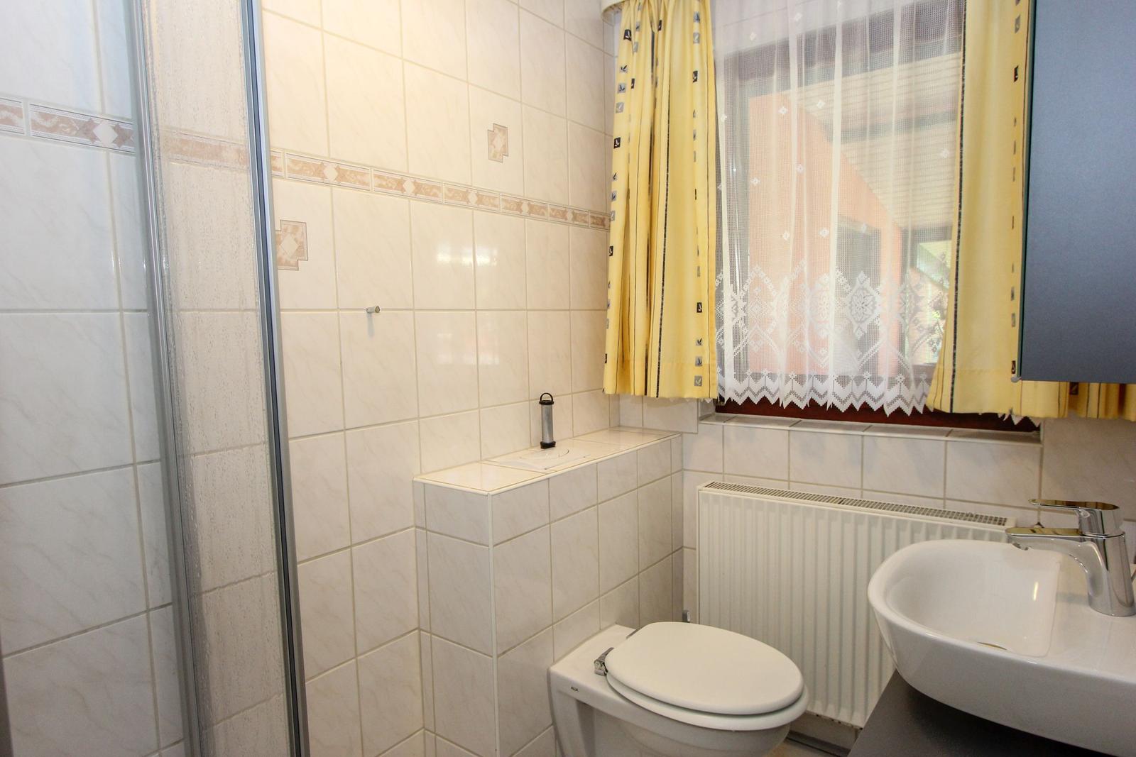 Badezimmer mit Dusche, Toilette und Waschbecken. Fenster mit gelben Vorhängen.