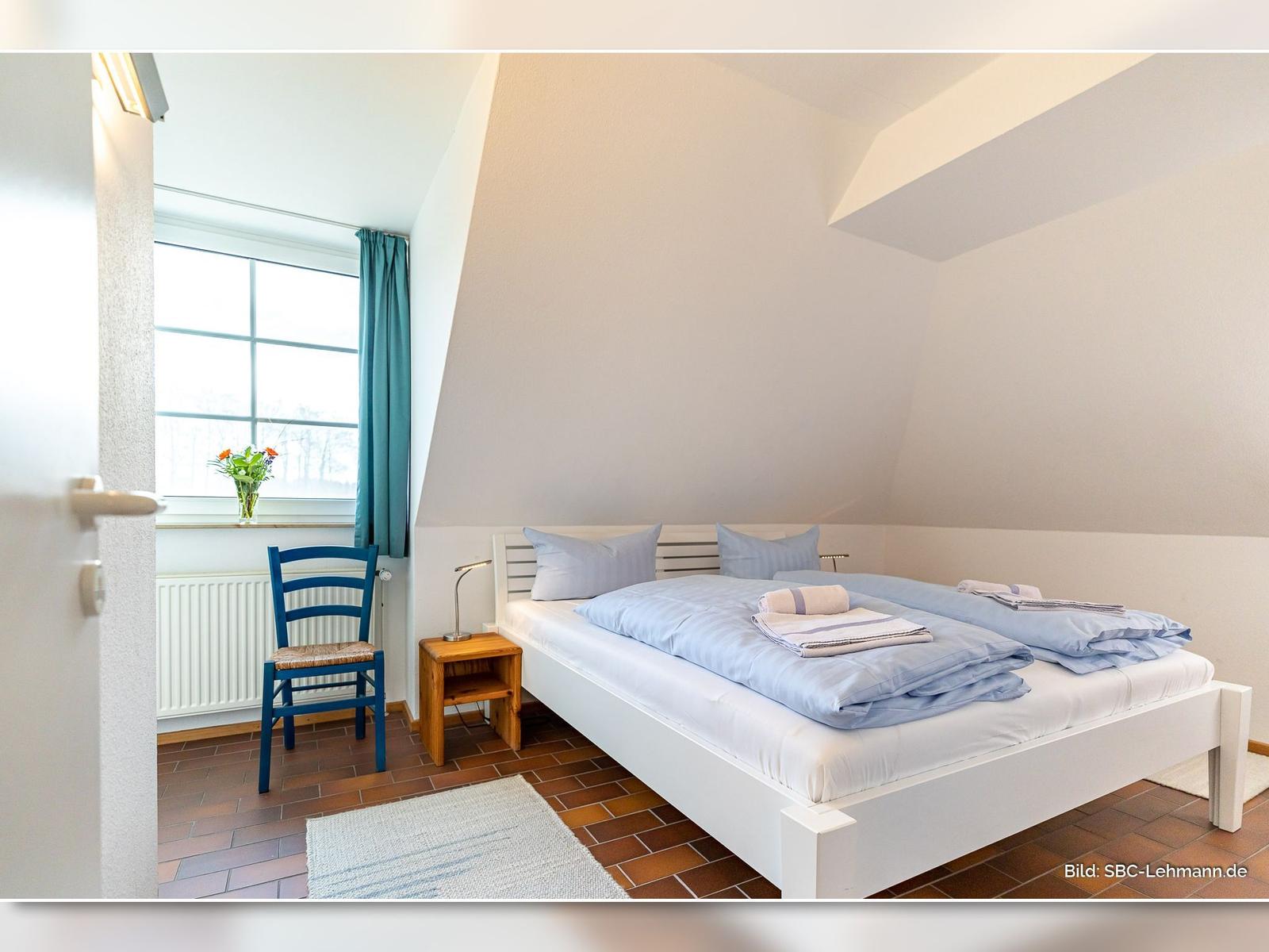 Schlafzimmer mit Doppelbett, Fenster, blauer Stuhl und Holzsideboard.