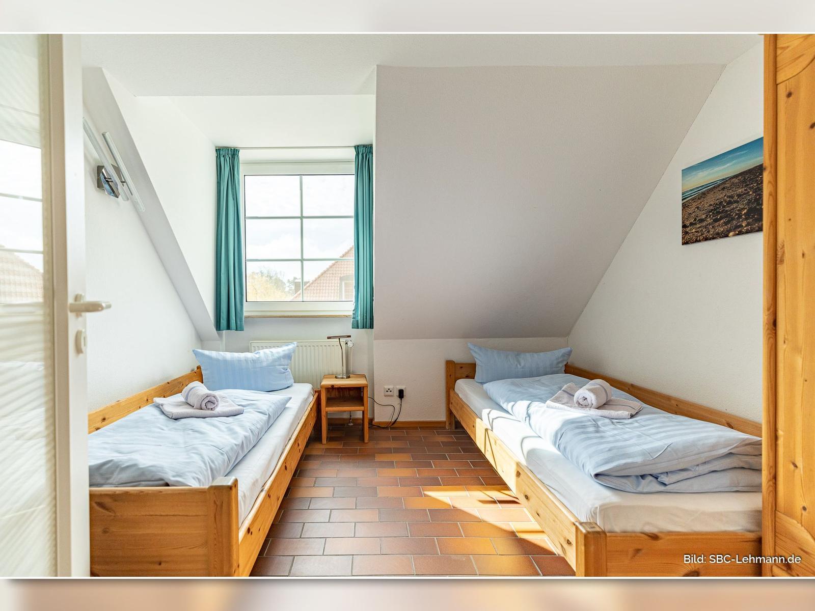 Zwei Einzelbetten mit blauen Bettwäschen, Holzrahmen und Fenster mit grünen Vorhängen.