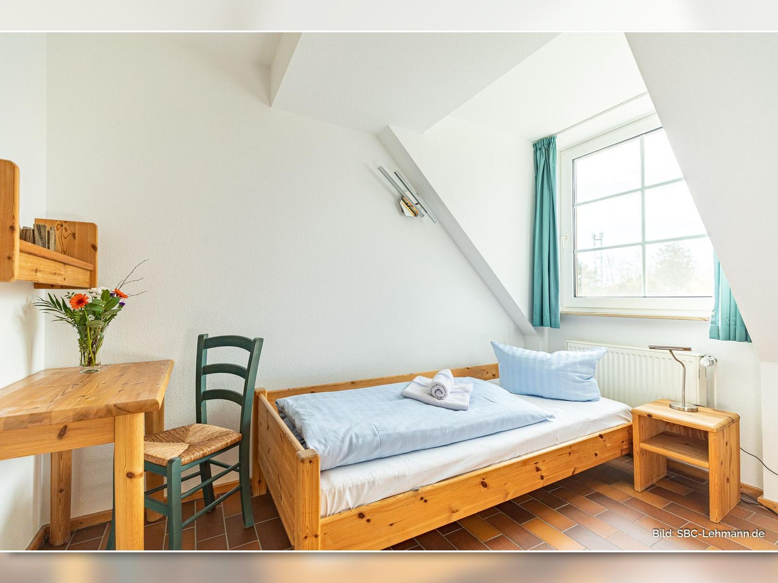 Ein Schlafzimmer mit Holzbett, Tisch, Stuhl und Fenster mit Vorhang.