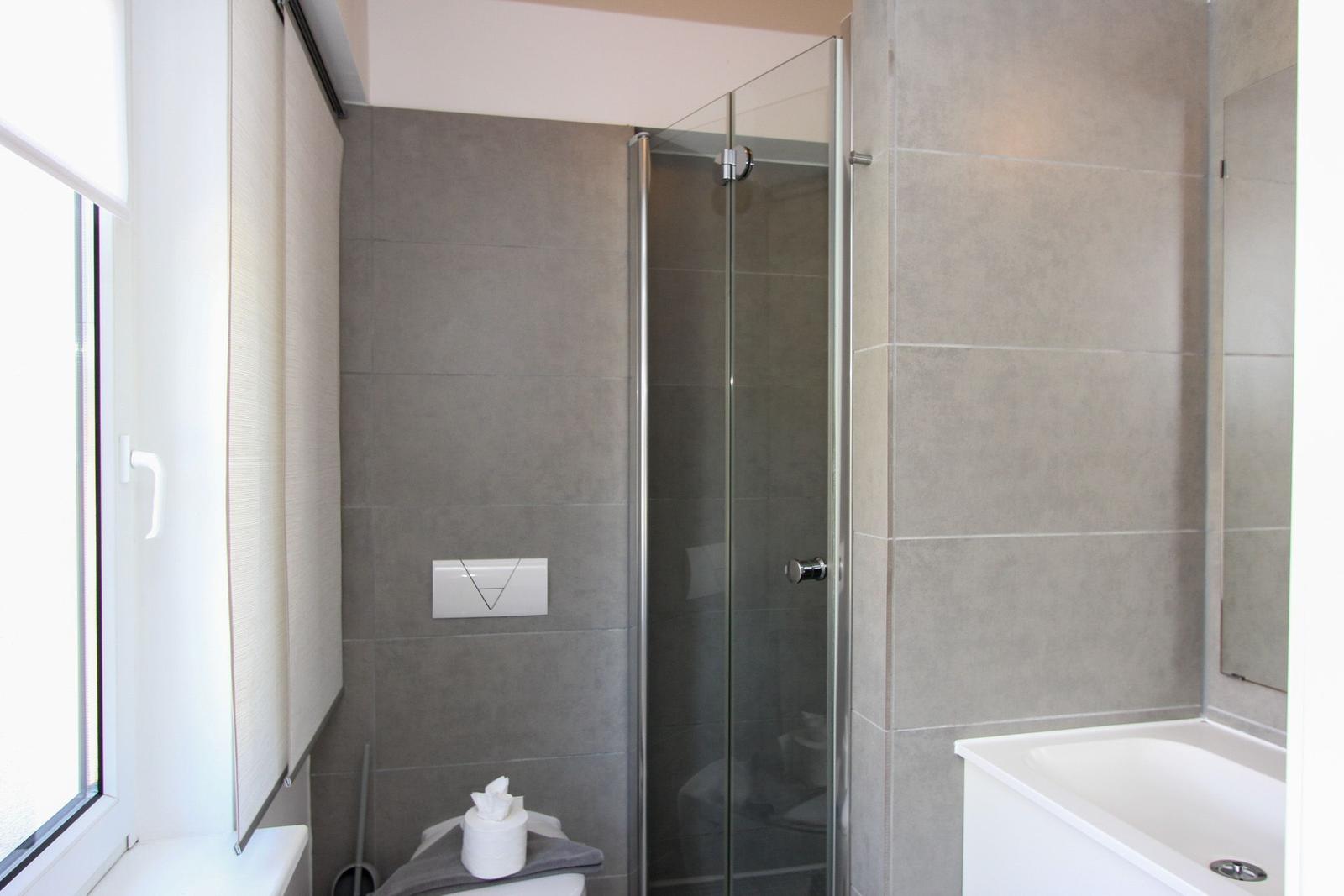Bad mit Dusche, Waschbecken und Fenster. Graue Fliesen und weiße Sanitärgeräte.