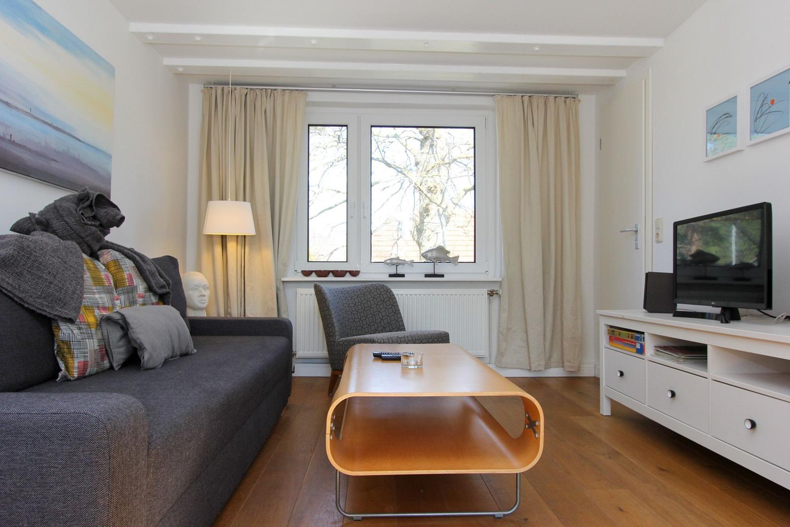 Wohnzimmer mit Sofa, Couchtisch und Fernseher. Fenster mit Vorhängen und Holzboden.
