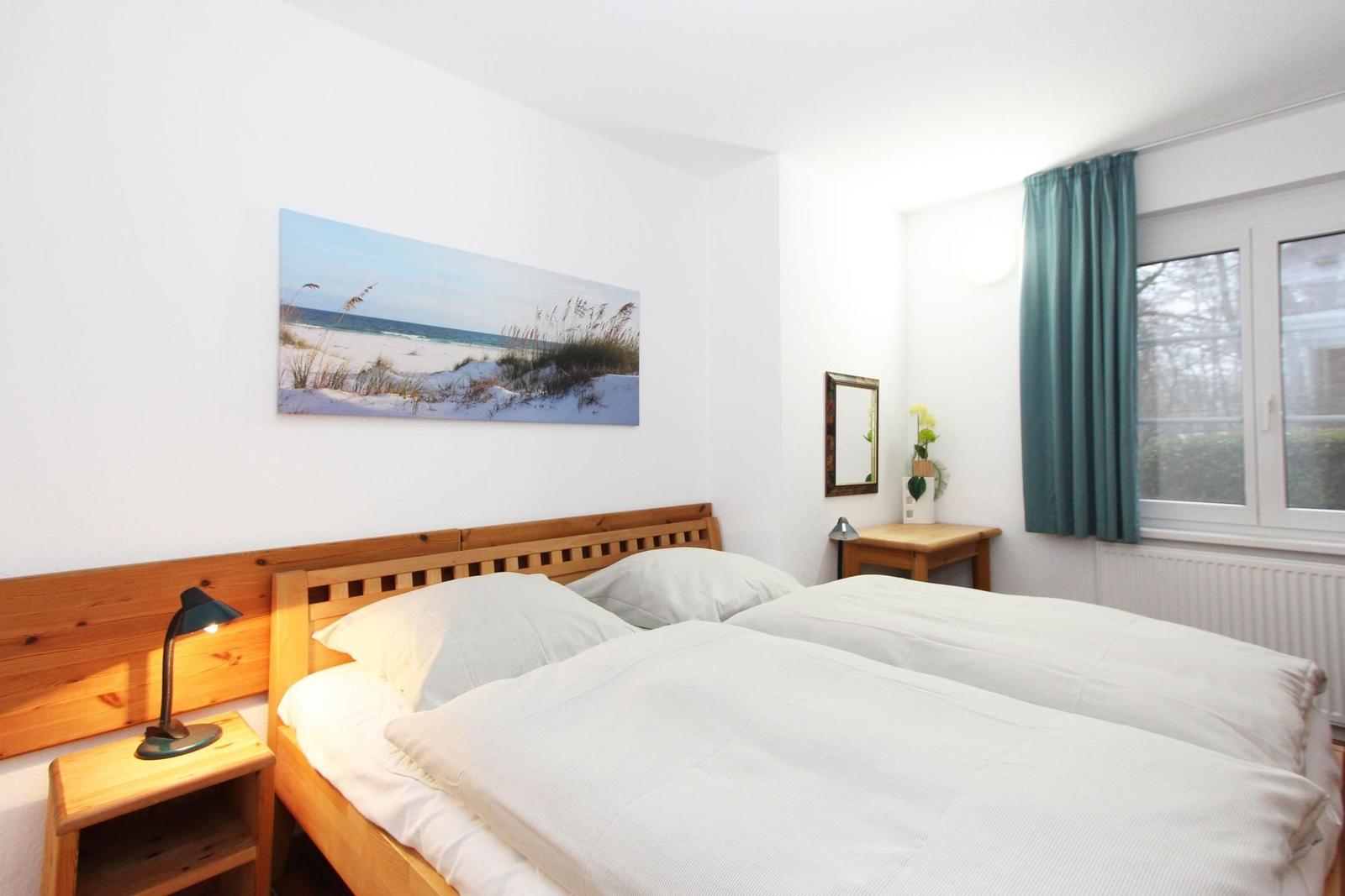 Schlafzimmer mit Doppelbett, Holzbett, Nachttisch und Fenster mit Vorhang.
