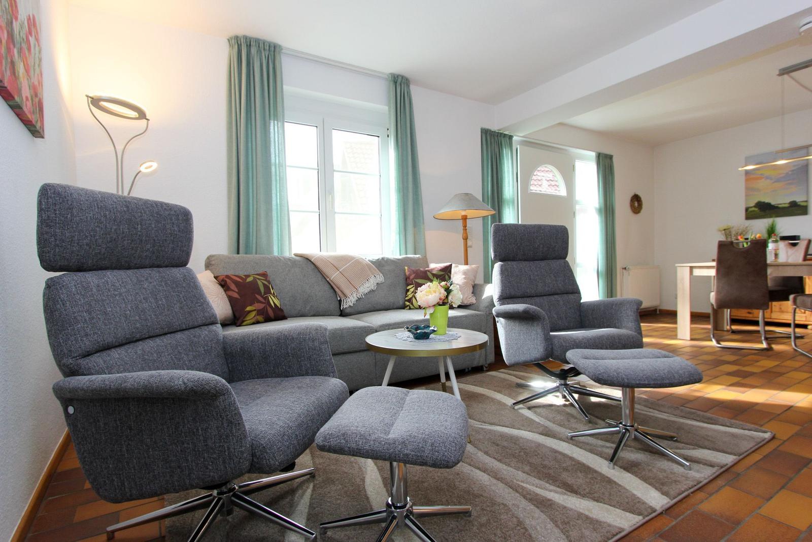 Wohnzimmer mit grauen Sesseln, Sofa und Tisch. Fenster mit grünen Vorhängen. Teppich und Holzboden.
