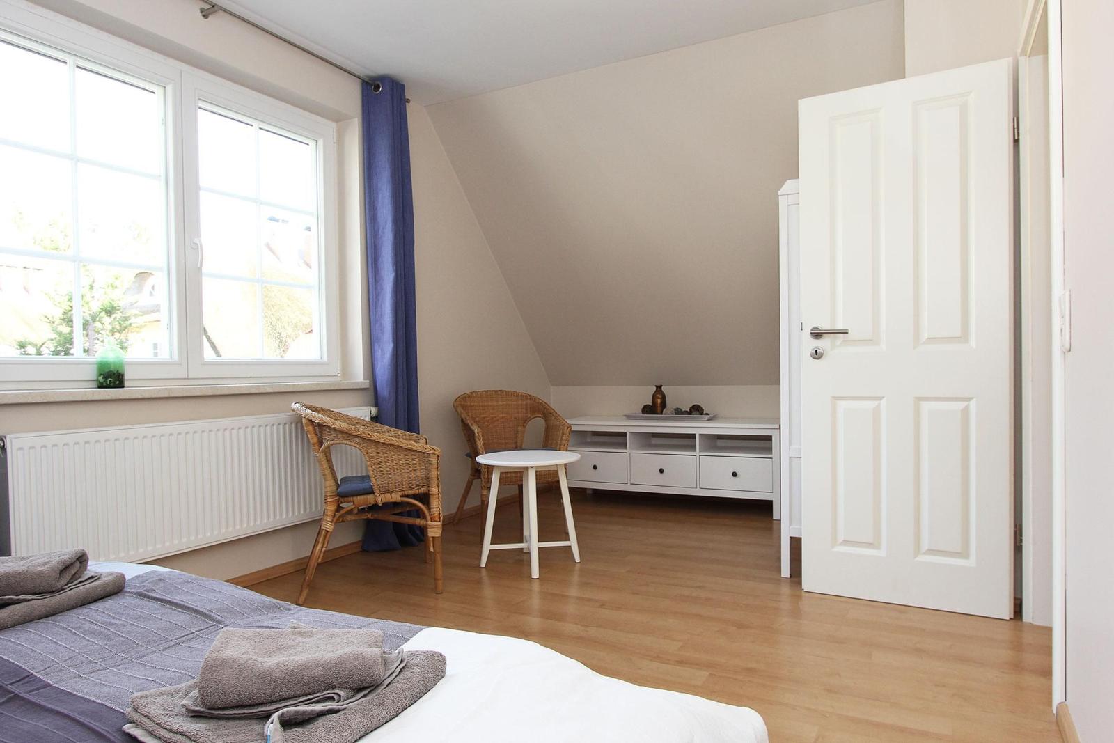 Ein Schlafzimmer mit Bett, Stuhl, Tisch und Fenster. Holzfußboden und weiße Wände.