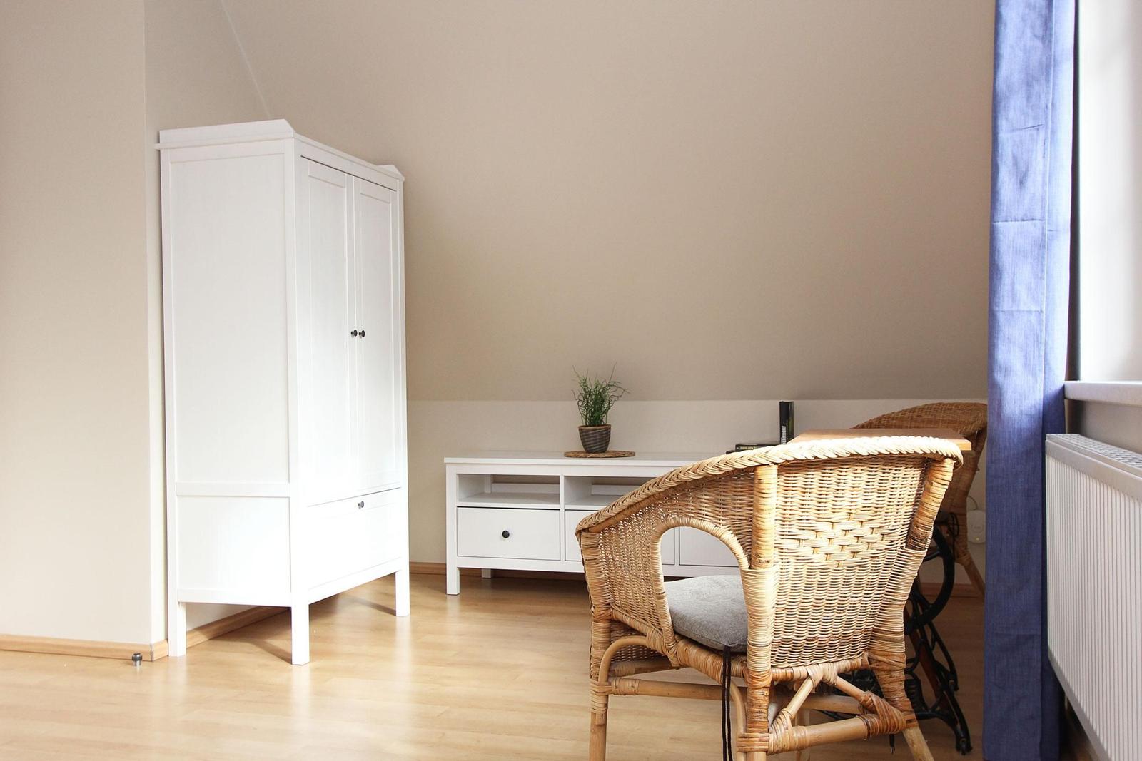 Ein Raum mit weißem Schrank, Wickerstuhl und Pflanze auf einem Tisch.
