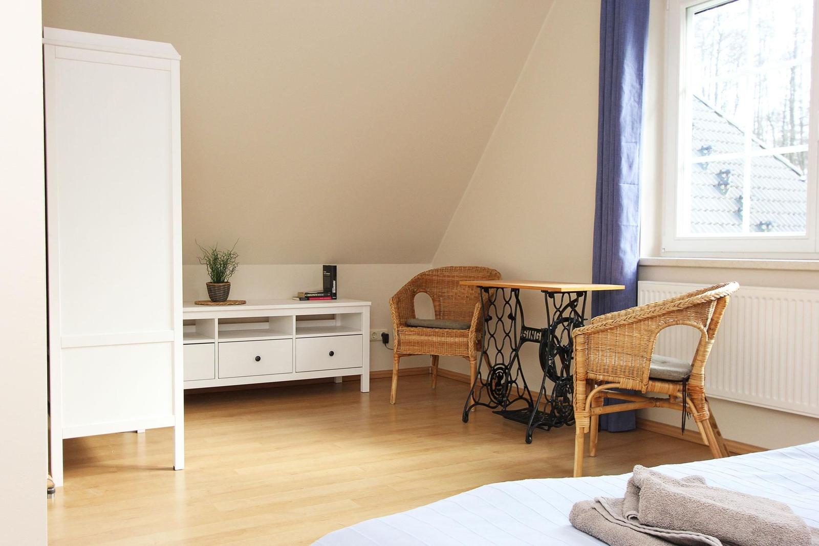 Zimmer mit Bett, Schrank, Tisch und zwei Stühlen. Fenster mit Vorhang. Holzboden.