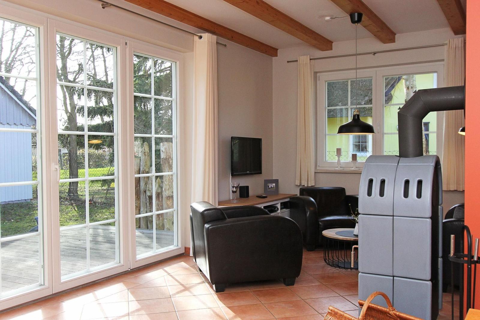 Wohnzimmer mit Sitzgelegenheiten, Fernseher und Kaminofen. Große Fenster mit Blick auf Garten.