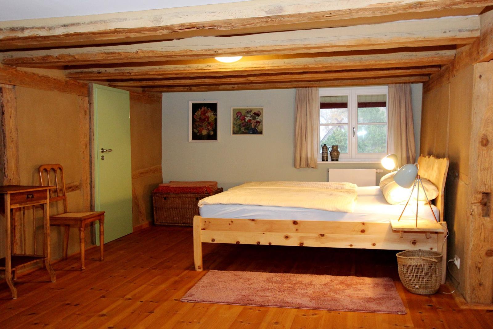 Schlafzimmer mit Holzbalken, Bett, Schreibtisch und Fenster.