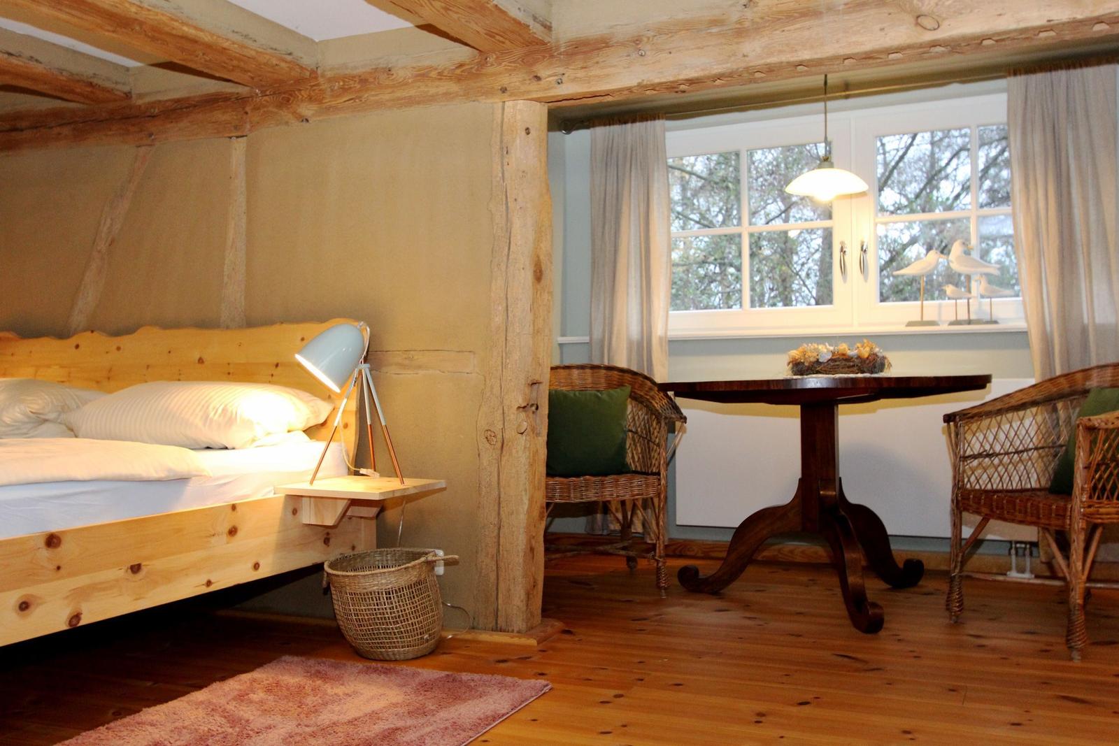 Schlafzimmer mit Holzbett, Tisch und Stühlen. Holzdecken und Boden. Fenster mit Vorhängen.