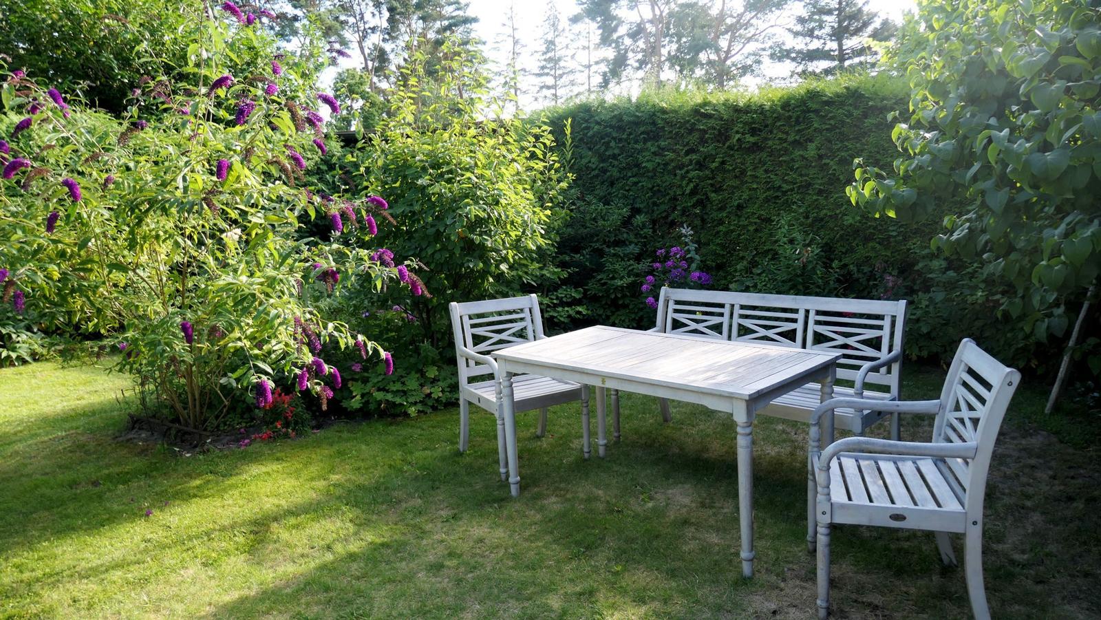 Helle Gartenmöbel auf Rasenfläche mit Blumenhecke und Bäumen im Hintergrund.
