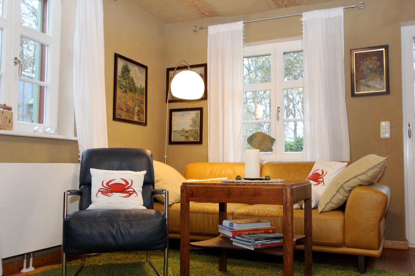 Ein Wohnzimmer mit gelbem Sofa, schwarzer Ledersessel und Holztisch. Fenster mit weißen Vorhängen.
