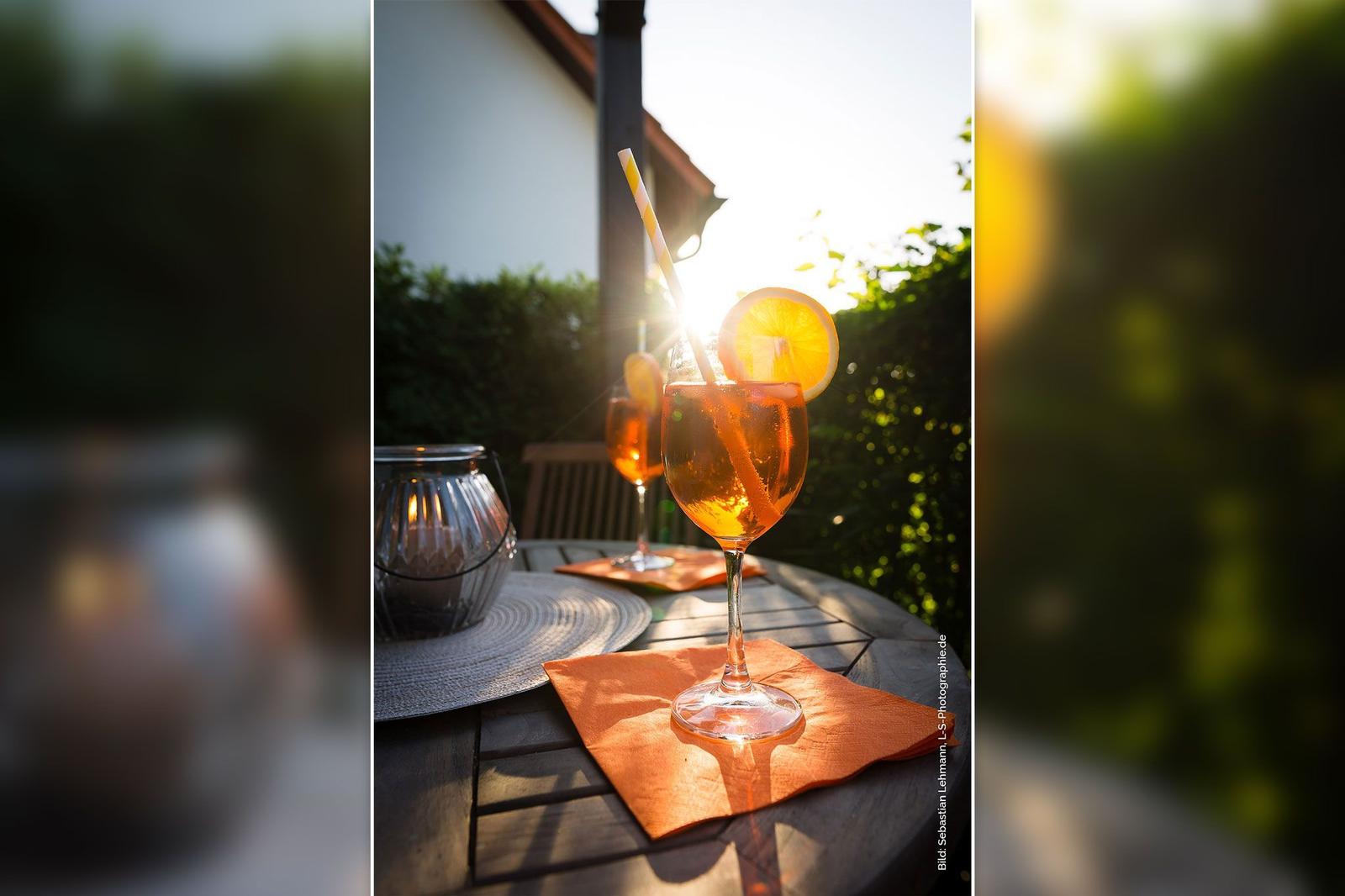 Ein Glas Aperol Spritz auf einer Terrasse mit Sonnenlicht und grüner Umgebung.