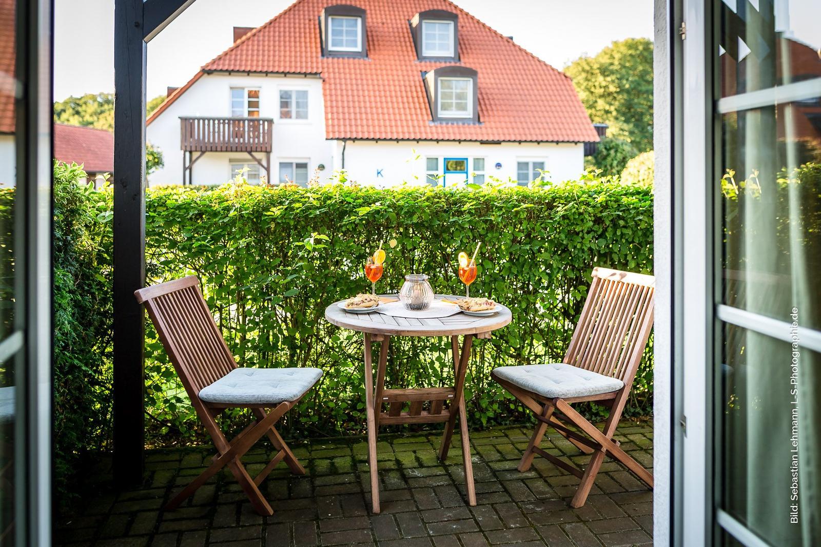 Terrasse mit Tisch und zwei Stühlen, dahinter grüner Heckenzaun und weißes Haus mit rotem Dach.