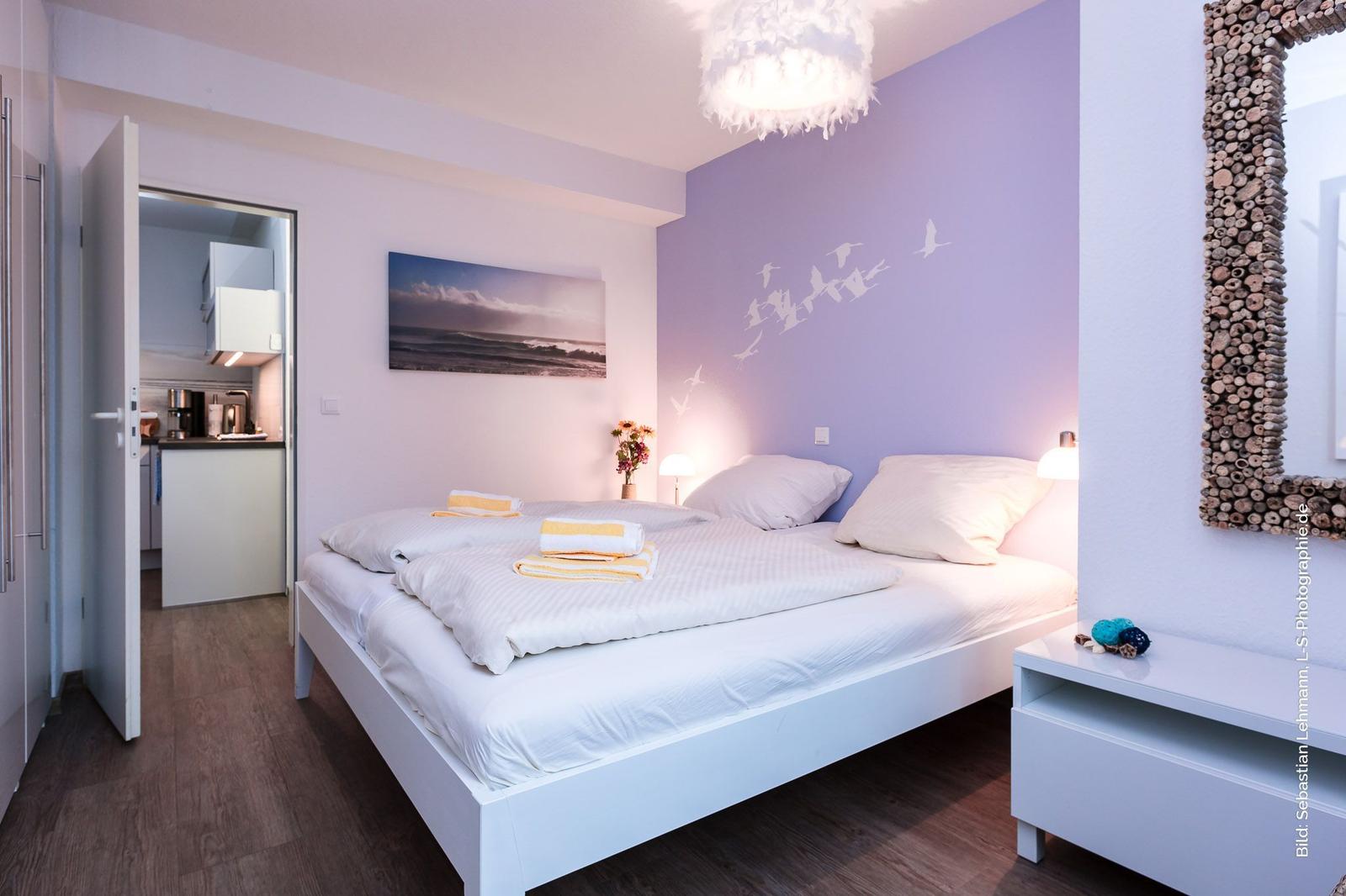 Schlafzimmer mit Bett, Wandbild und Spiegel. Durchgang zur Küche sichtbar.