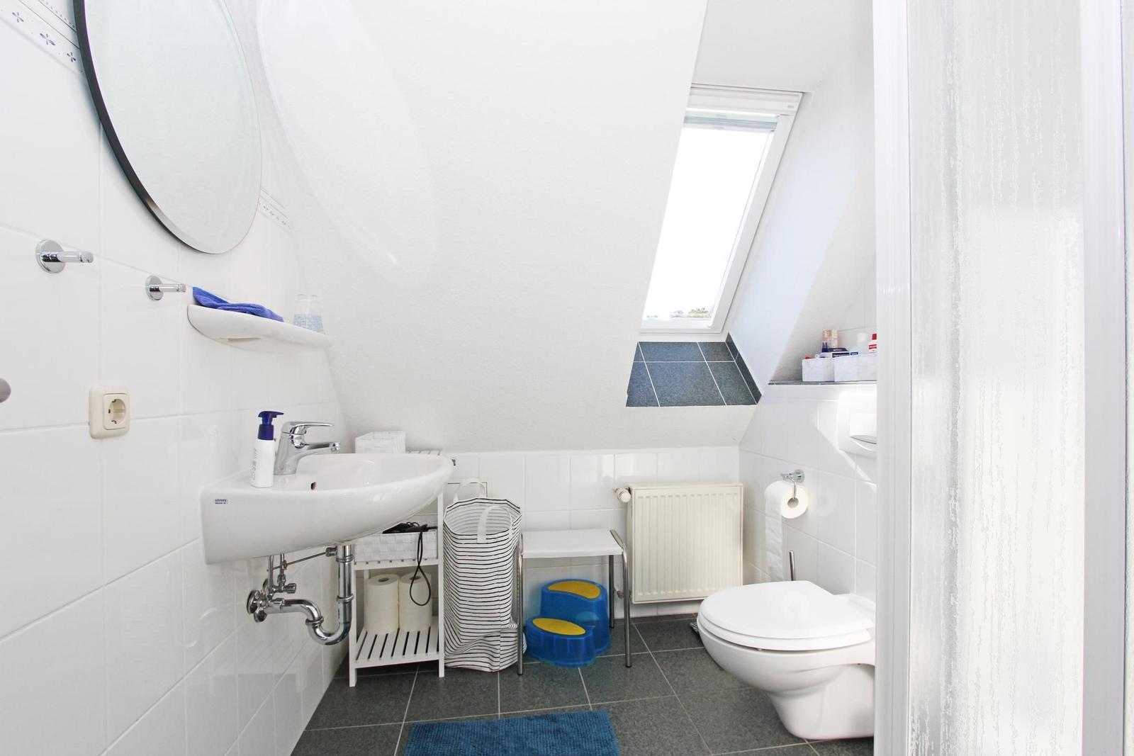 Badezimmer mit Waschbecken, Toilette und Dachfenster. Weiße Fliesen und graue Bodenfliesen.