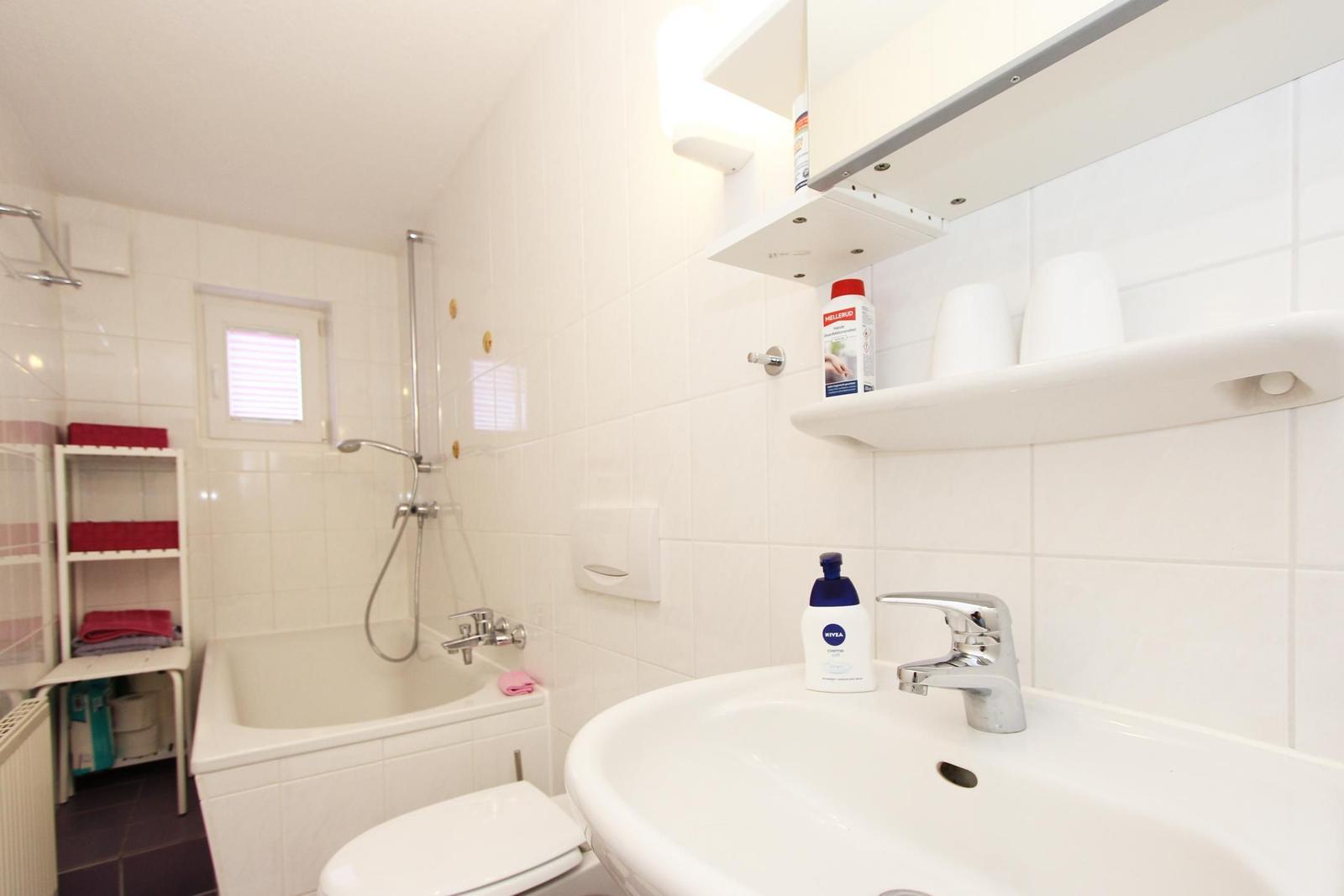 Badezimmer mit Badewanne, Dusche, Waschbecken und Toilette. Weiße Fliesen und Regale.