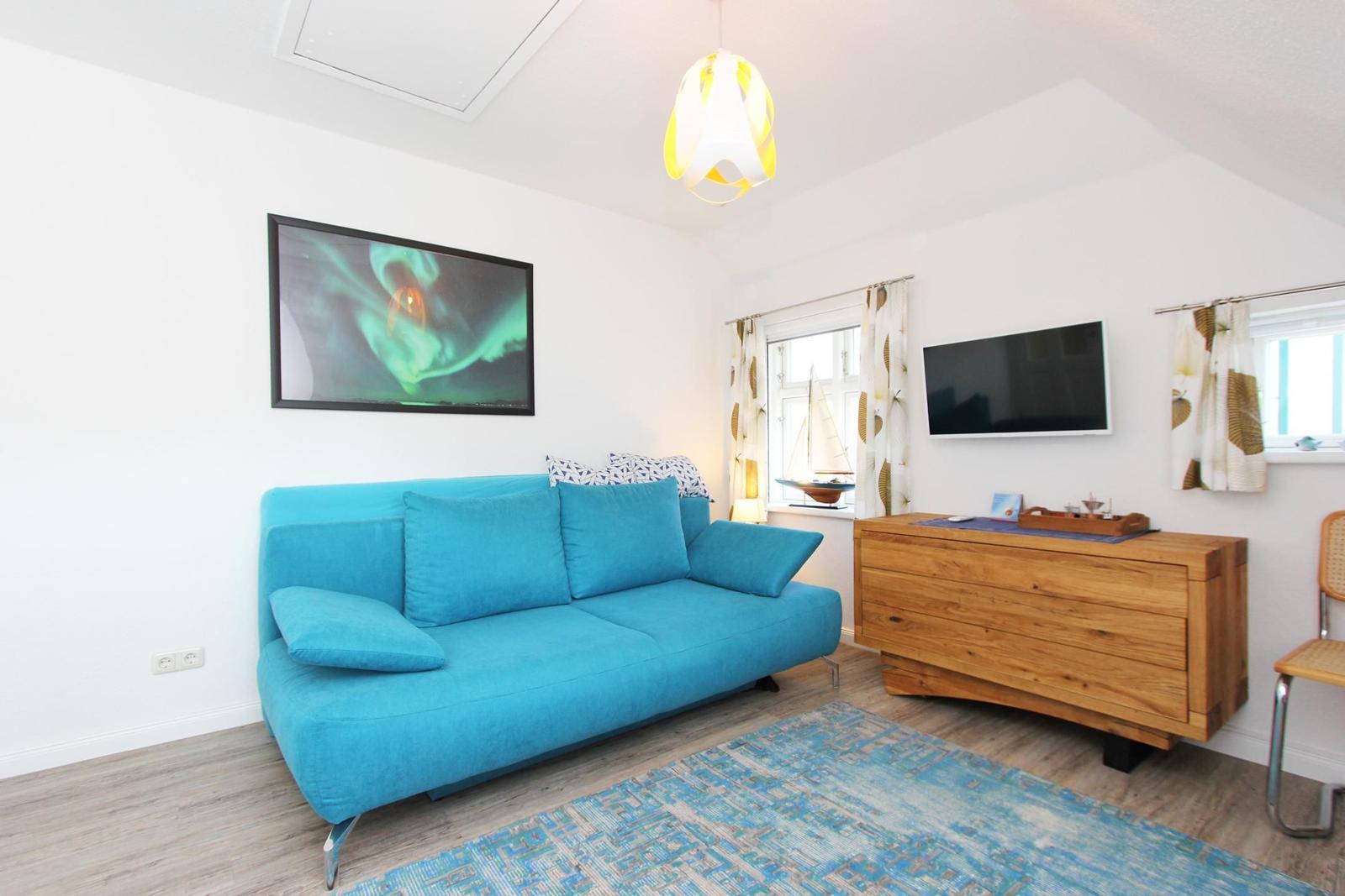 Ein Wohnzimmer mit blauem Sofa, Holzschrank und Wandbild. Fenster mit Vorhängen.