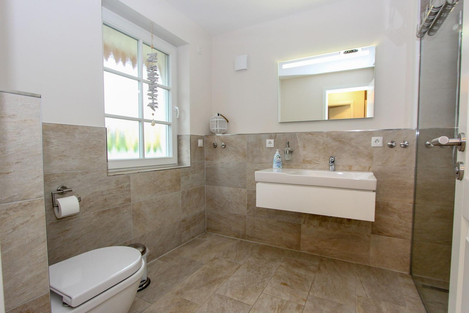 Badezimmer mit Toilette, Waschbecken und Dusche. Fenster mit Vorhang und Spiegel über Waschbecken.