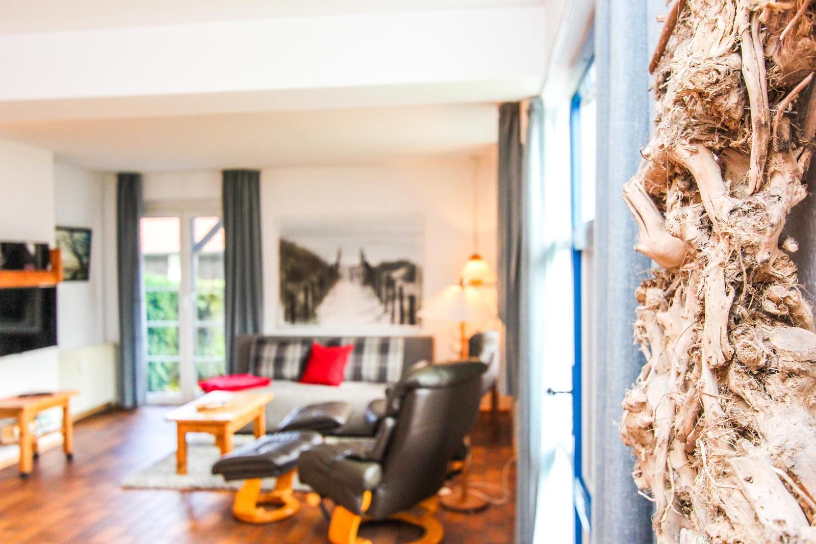 Wohnzimmer mit Sofa, Sessel und Holztisch. Fenster mit Blick auf Garten. Dekorative Baumrinde an Wand.