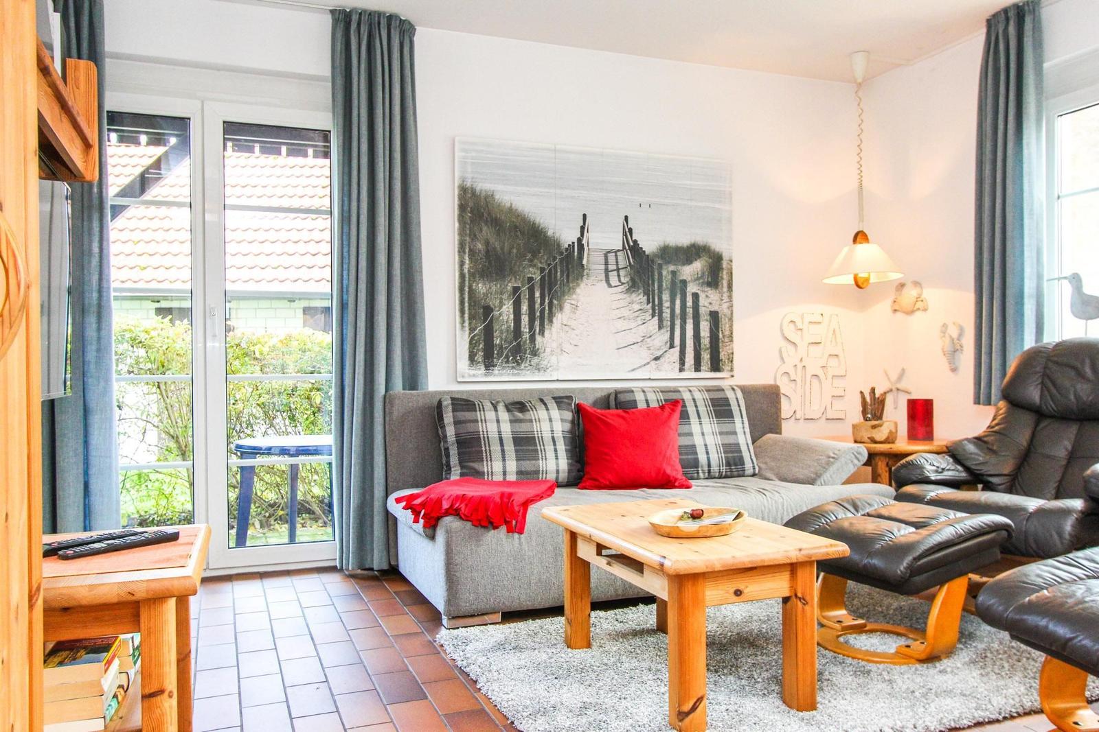 Wohnzimmer mit Sofa, Couchtisch und Sessel. Wandbild zeigt Strandweg. Fenster mit Blick auf Garten.