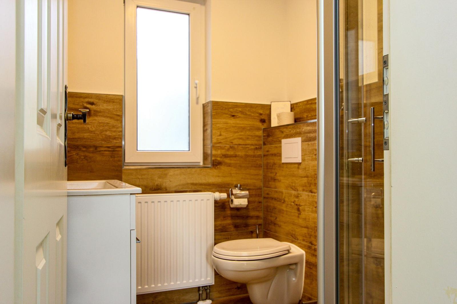Badezimmer mit Toilette, Dusche und Fenster. Holzwandverkleidung und weißer Waschtisch.