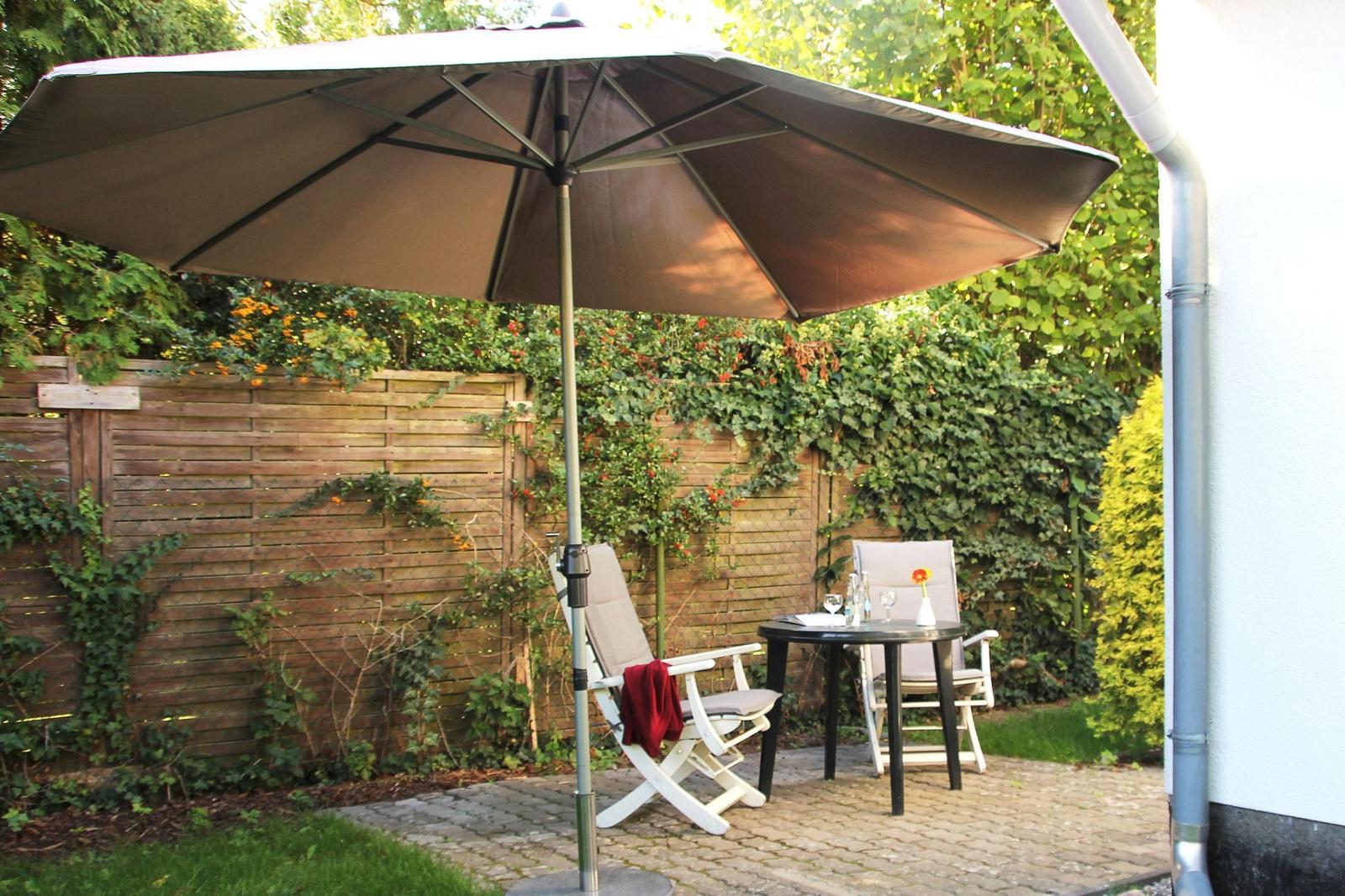 Terrasse mit Sonnenschirm, Tisch und zwei Stühlen. Hintergrund: Holzzaun und grüne Pflanzen.