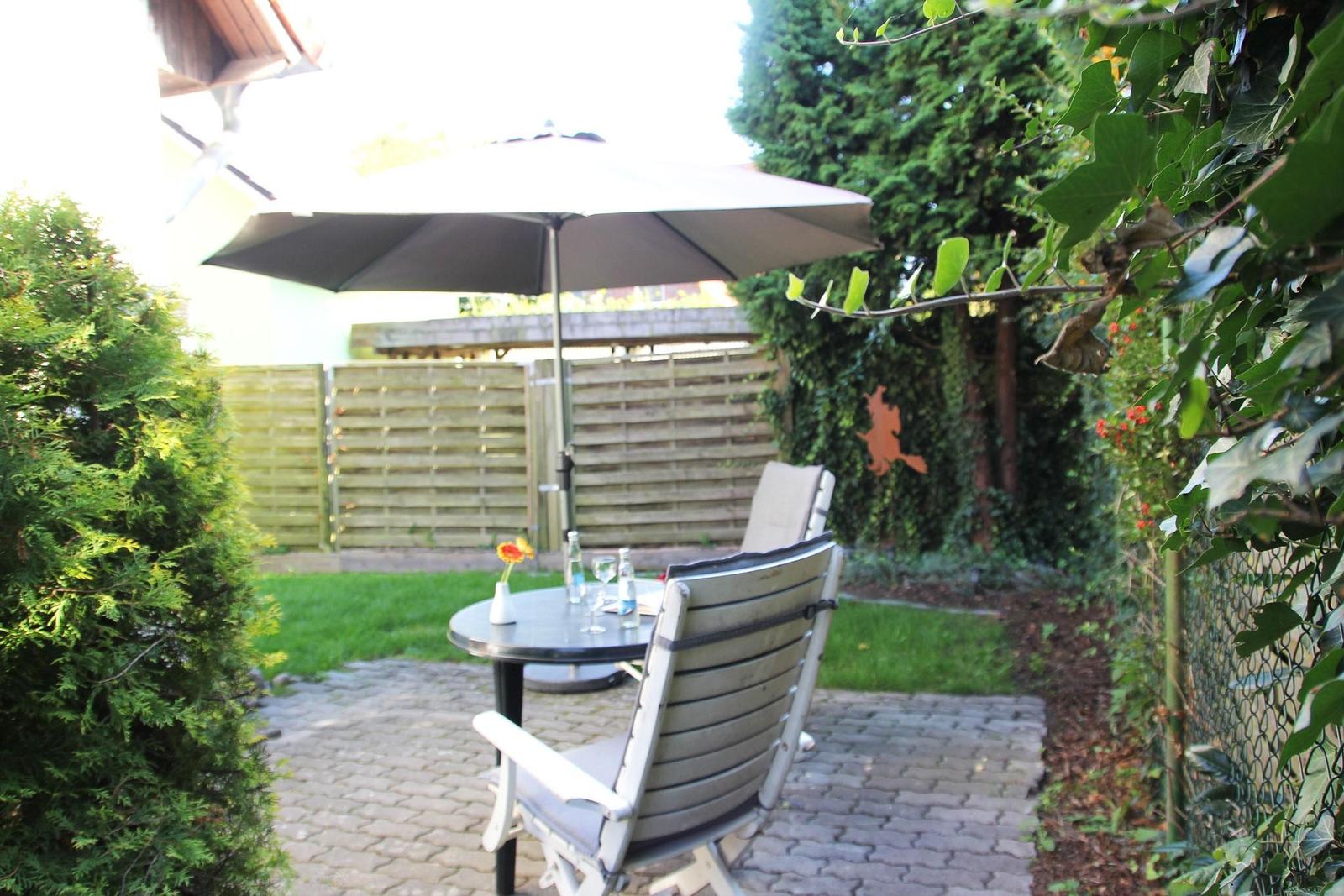 Hinterhof mit Tisch, Stühlen und Sonnenschirm. Grasfläche und Beete umgeben den Platz.
