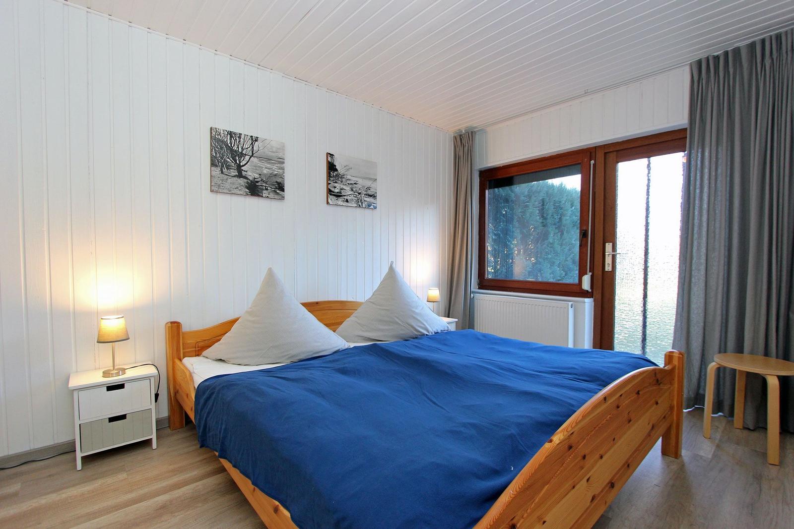 Schlafzimmer mit Holzbett, blauer Bettwäsche, Fenster mit Blick auf Wasser und Wald.