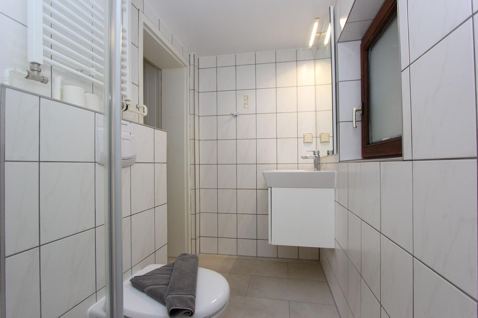 Badezimmer mit Dusche, Waschbecken und Toilette. Weiße Fliesen und Holzfenster.