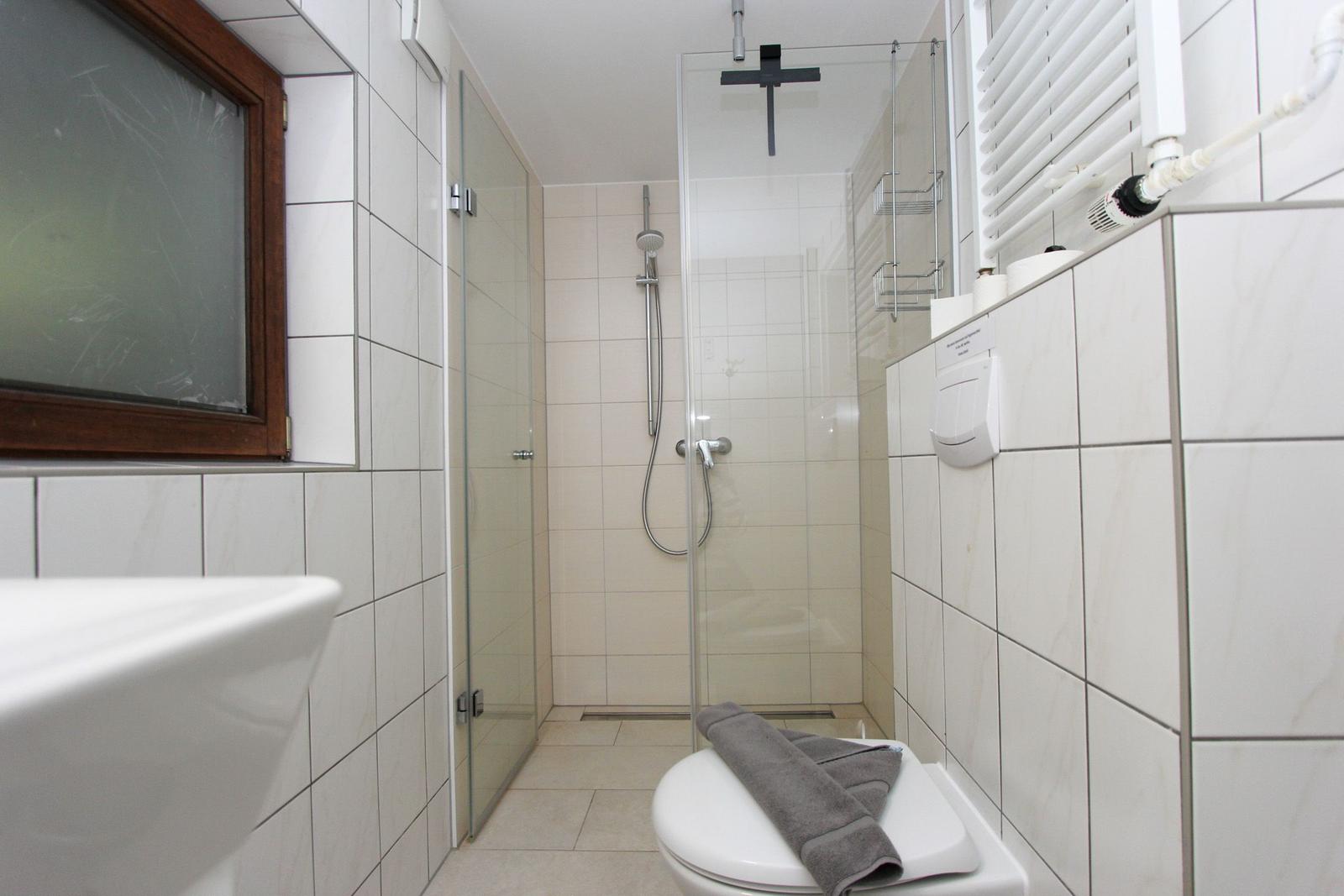 Badezimmer mit Dusche, Toilette und Waschbecken. Weiße Fliesen und Fenster mit Holzrahmen.