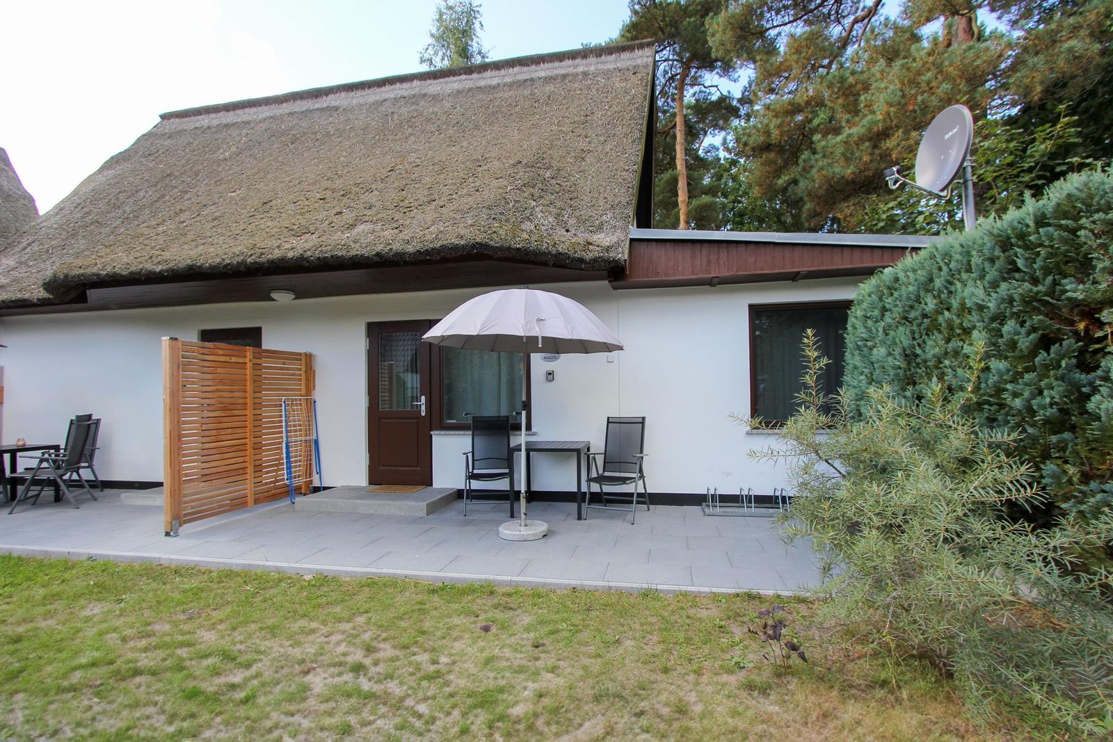 Haus mit strohgedecktem Dach, Terrasse mit Tisch, Stühlen und Sonnenschirm.
