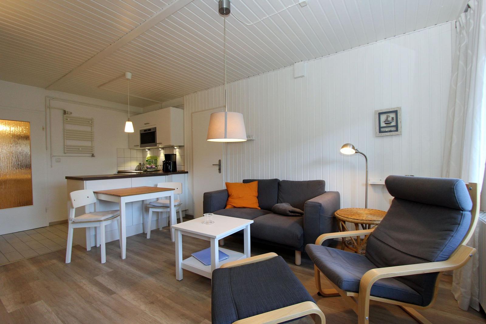 Wohnbereich mit Sofa, Sessel und Küchenbereich. Holzboden und weiße Wände.