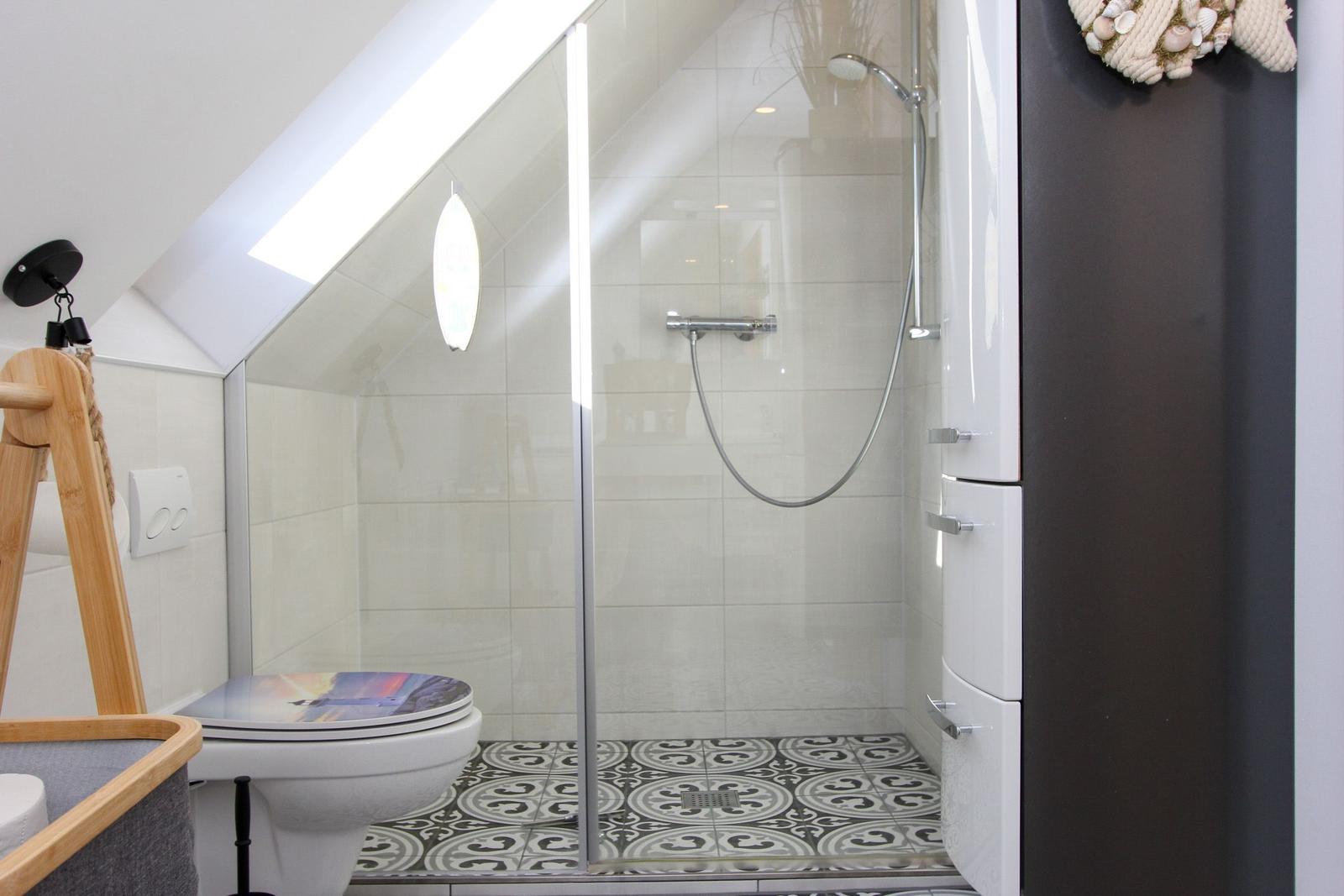 Dusche mit Glaswand, Toilette und schwarz-weißem Bodenfliesenmuster unter Dachschrägen.