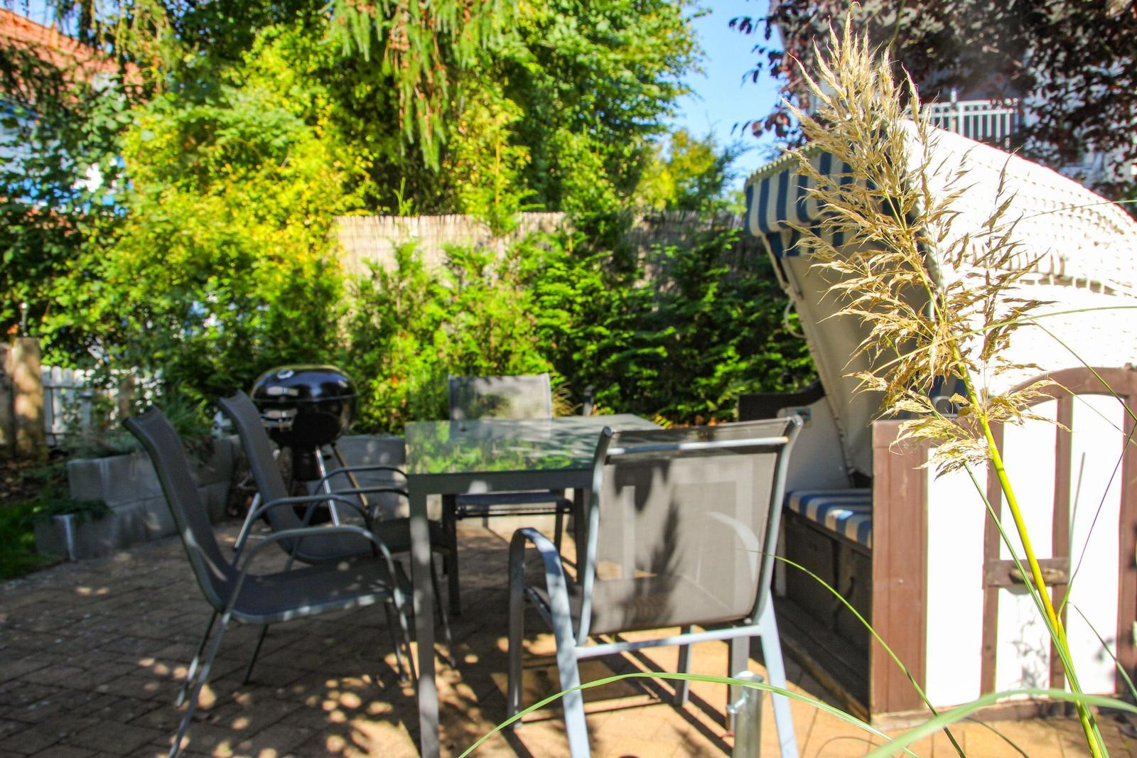 Terrasse mit Tisch, Stühlen, Grill und Strandkorb. Umgeben von grüner Vegetation.