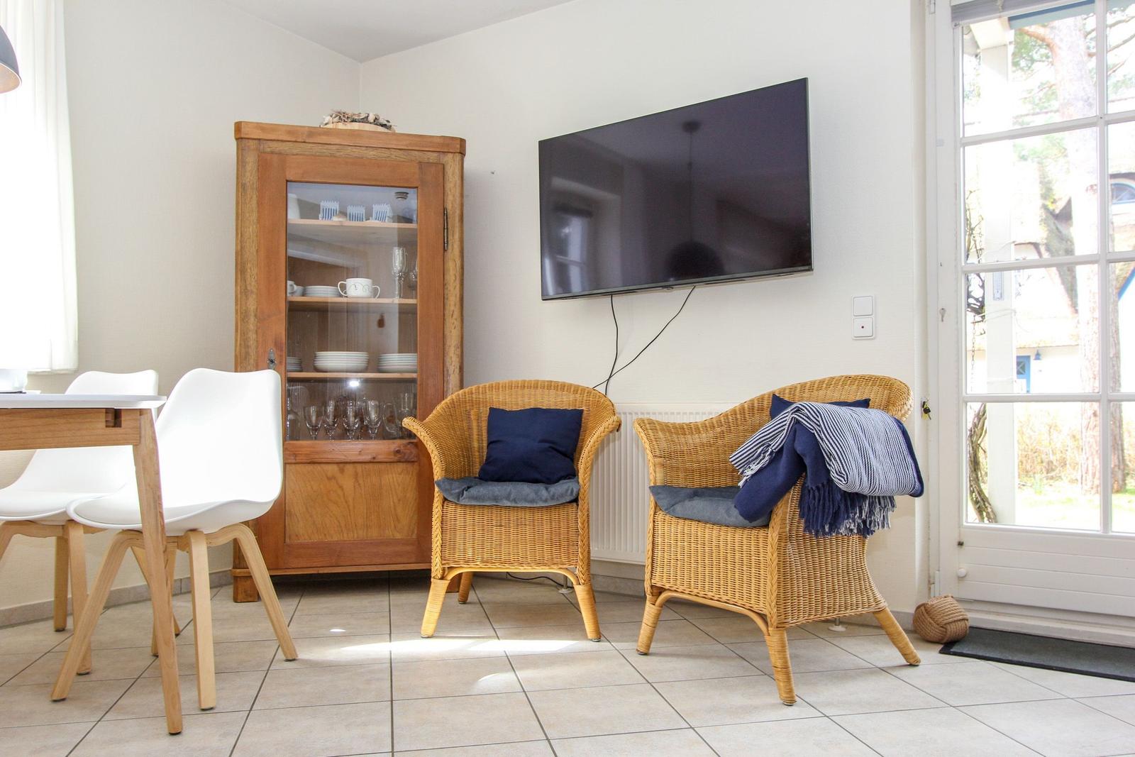 Zwei Rattan-Stühle mit Kissen, Fernseher und Essbereich mit Holztisch und weißen Stühlen.