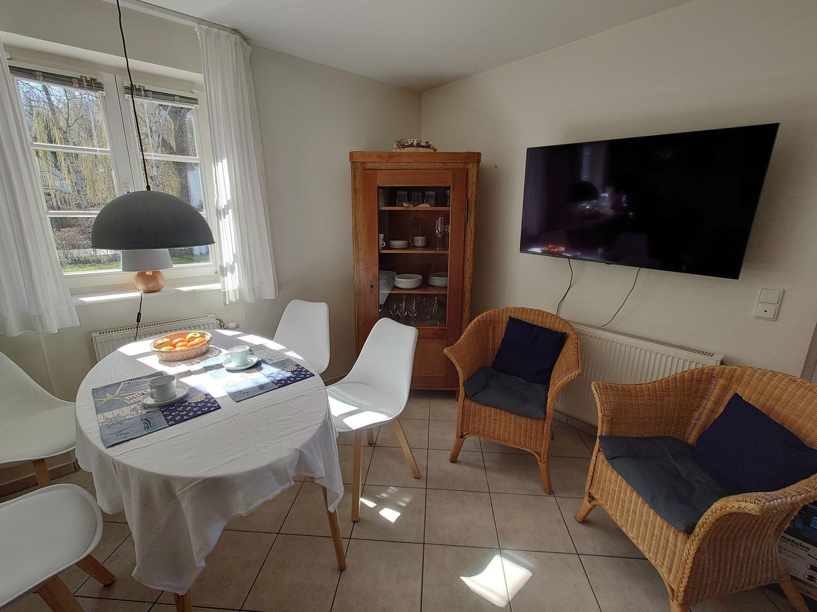 Essbereich mit runder Tisch, weißen Stühlen und TV-Wand. Rattan-Stühle mit Kissen.