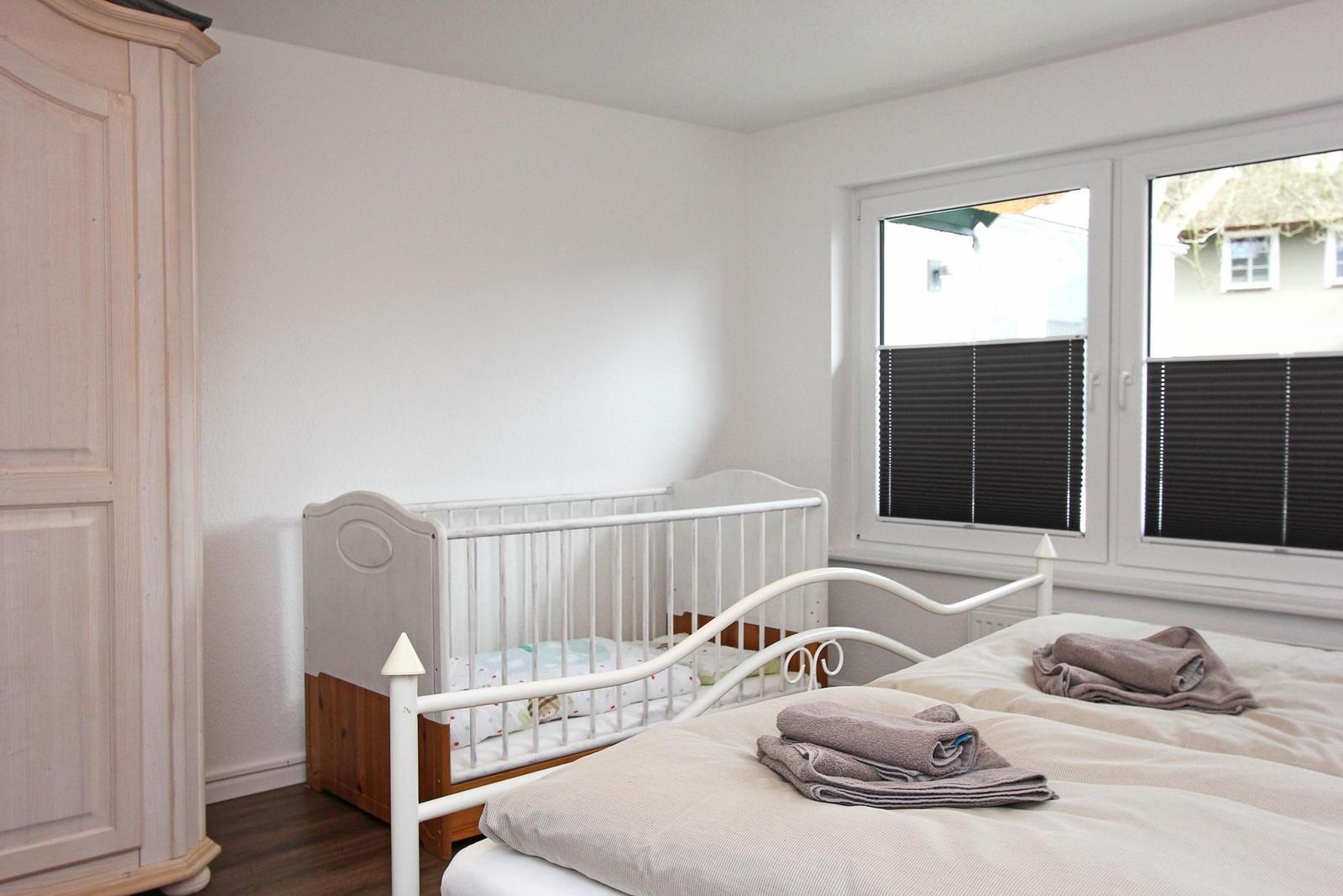 Zimmer mit Babybett, Bett und Fenster mit Jalousien.