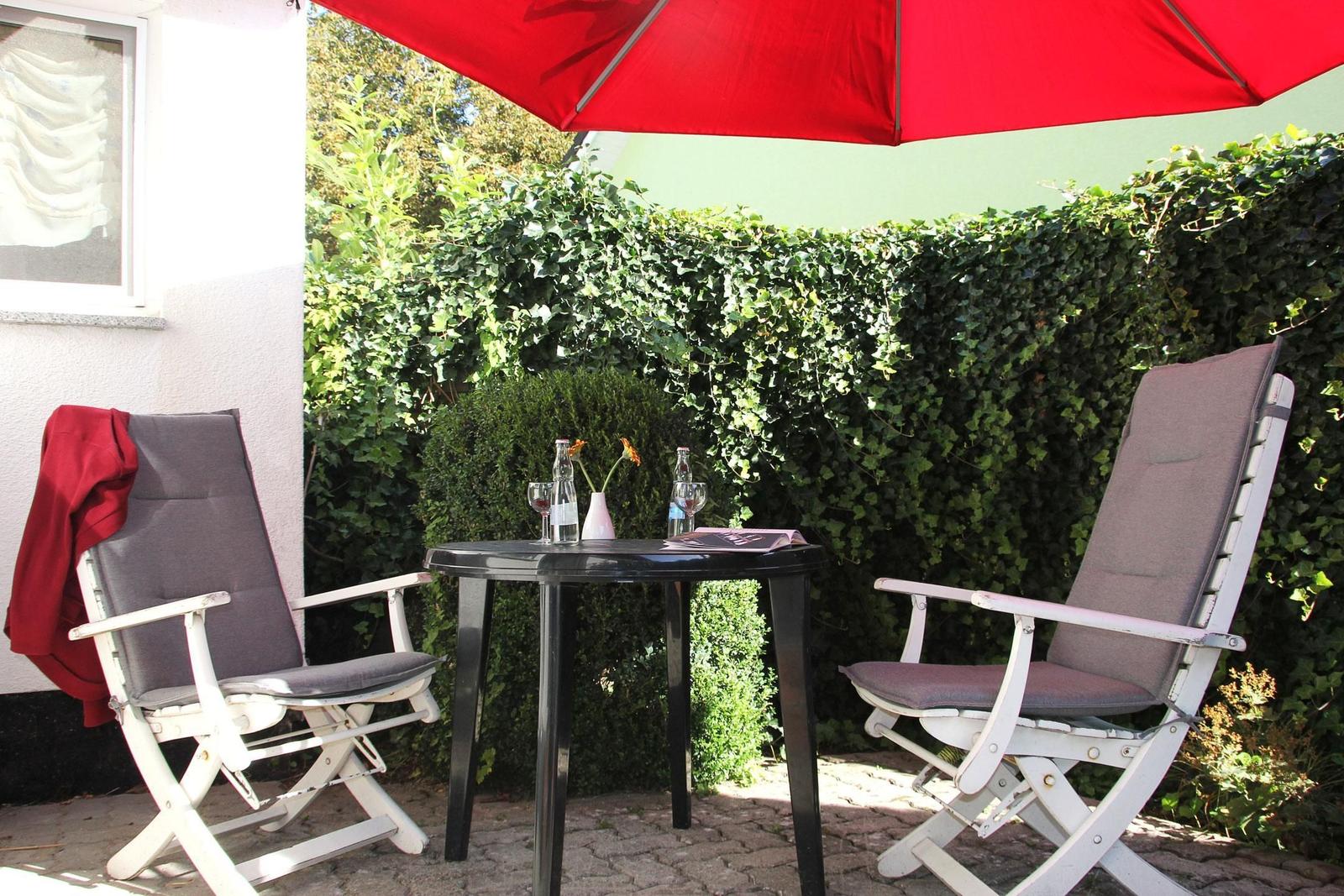 Terrasse mit Tisch, zwei Stühlen und rotem Sonnenschirm. Hintergrund: grüner Heckenzaun.
